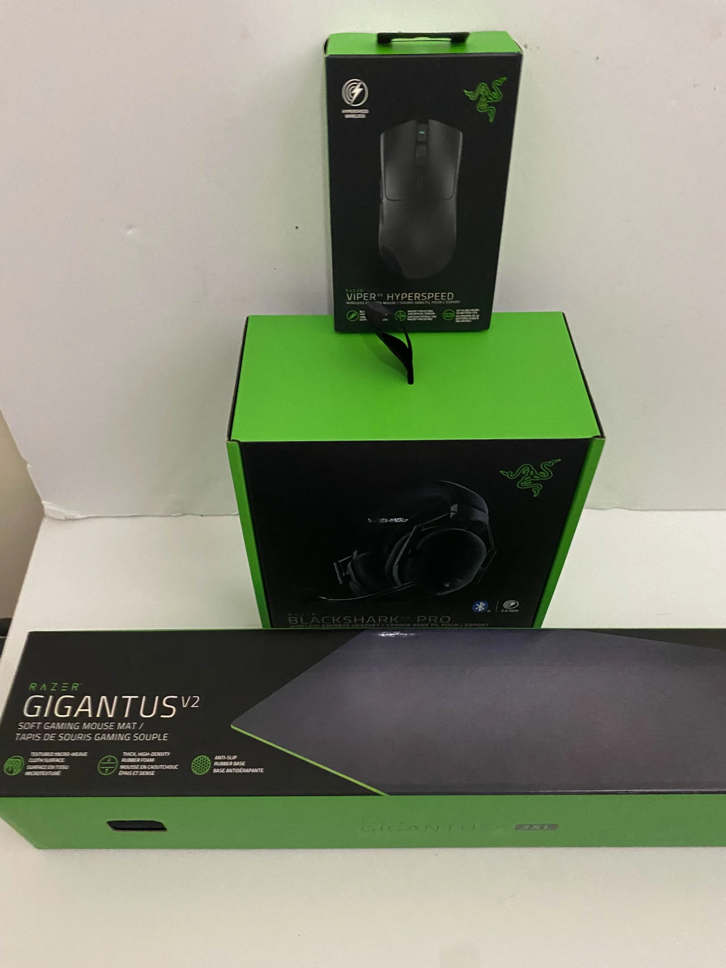 A gaming mouse, a gaming headset, and a gaming mouse pad, all in green and black packaging, placed on a white surface against a plain wall.