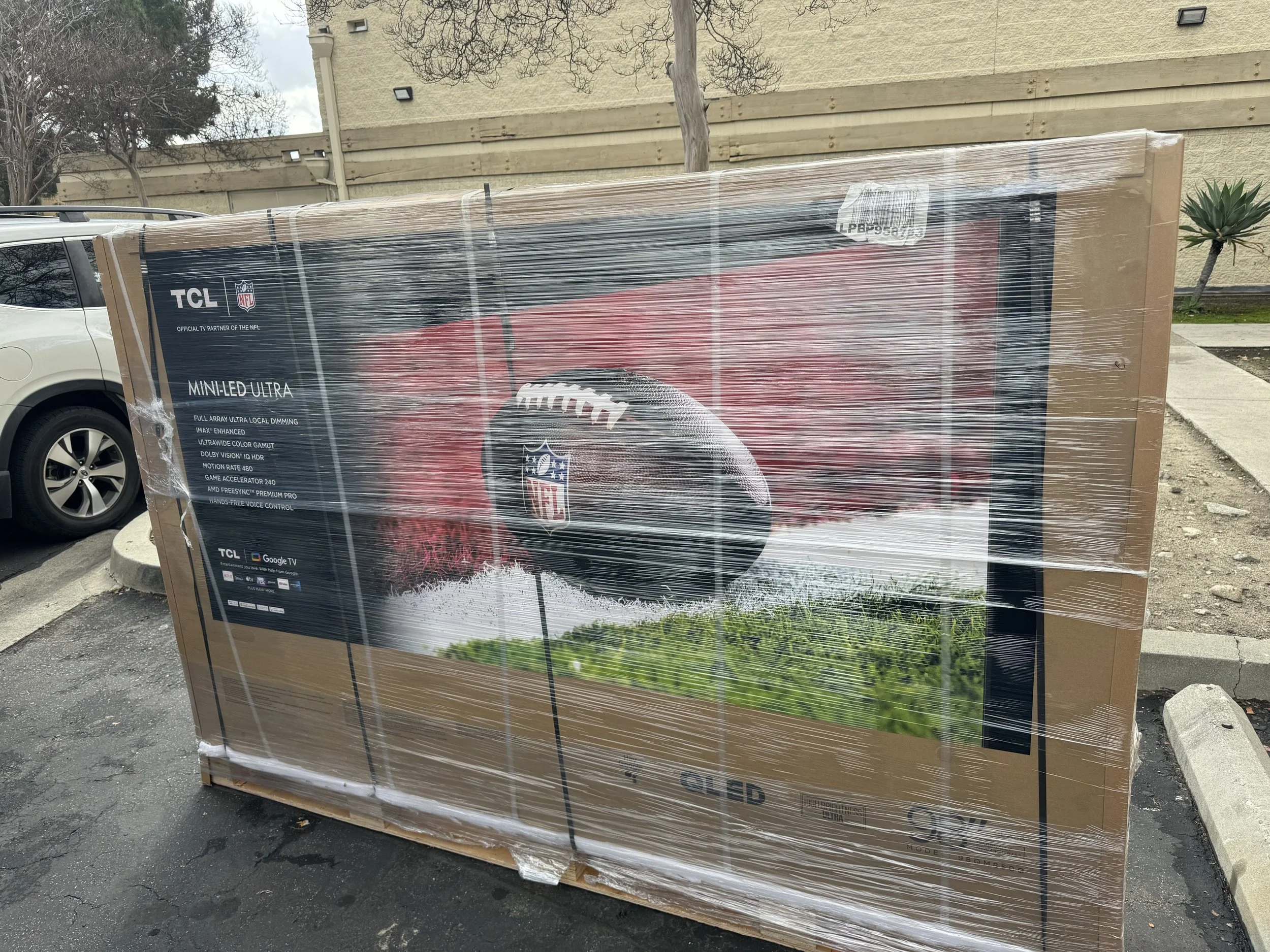 Unboxed TCL Mini LED Ultra TV in a parking lot, wrapped in plastic, with an NFL football logo on the screen.