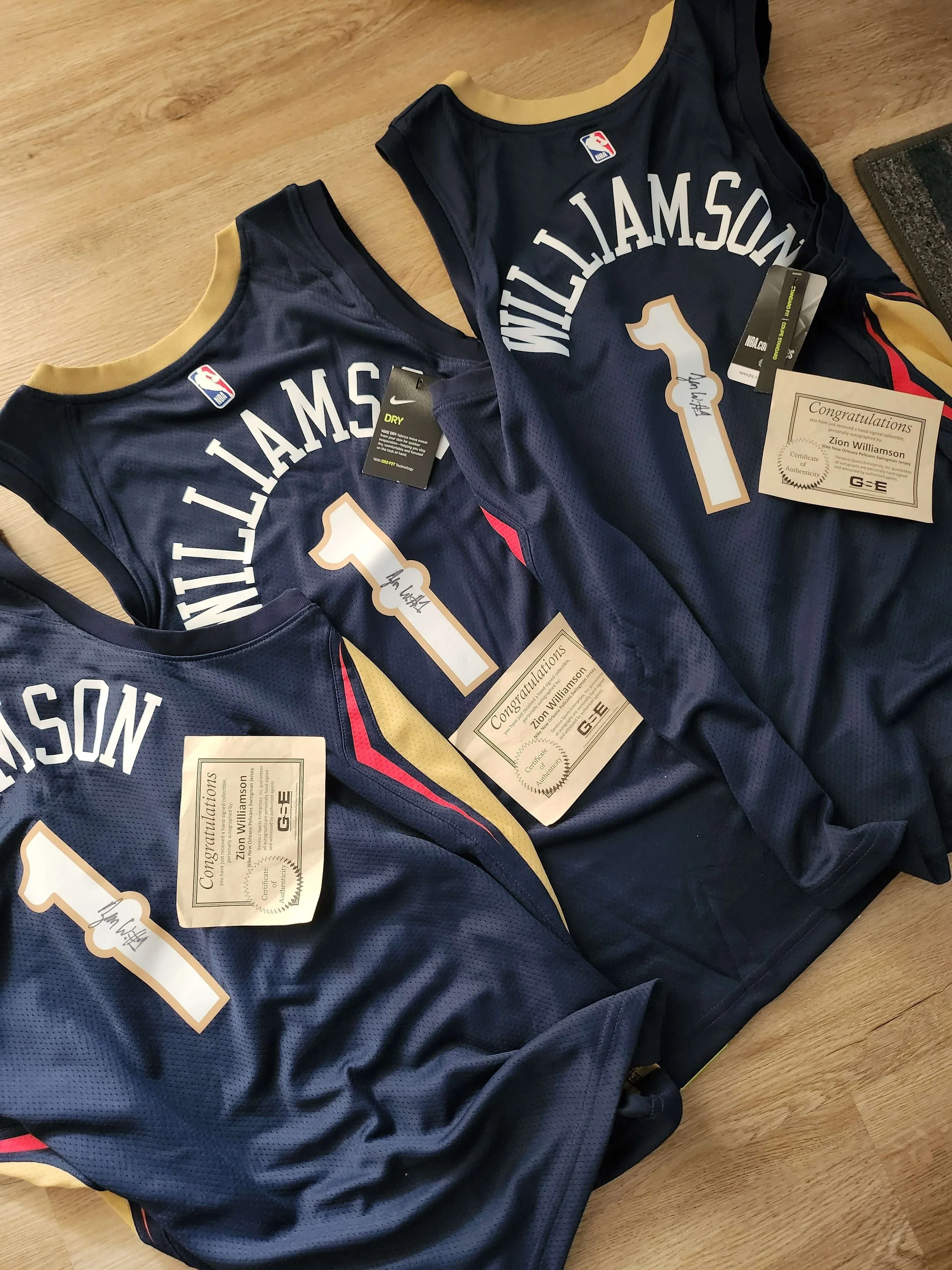 Three basketball jerseys with 'Willaimson' and the number 1 on the back, along with congratulatory certificates and signed paper, placed on a wooden floor.