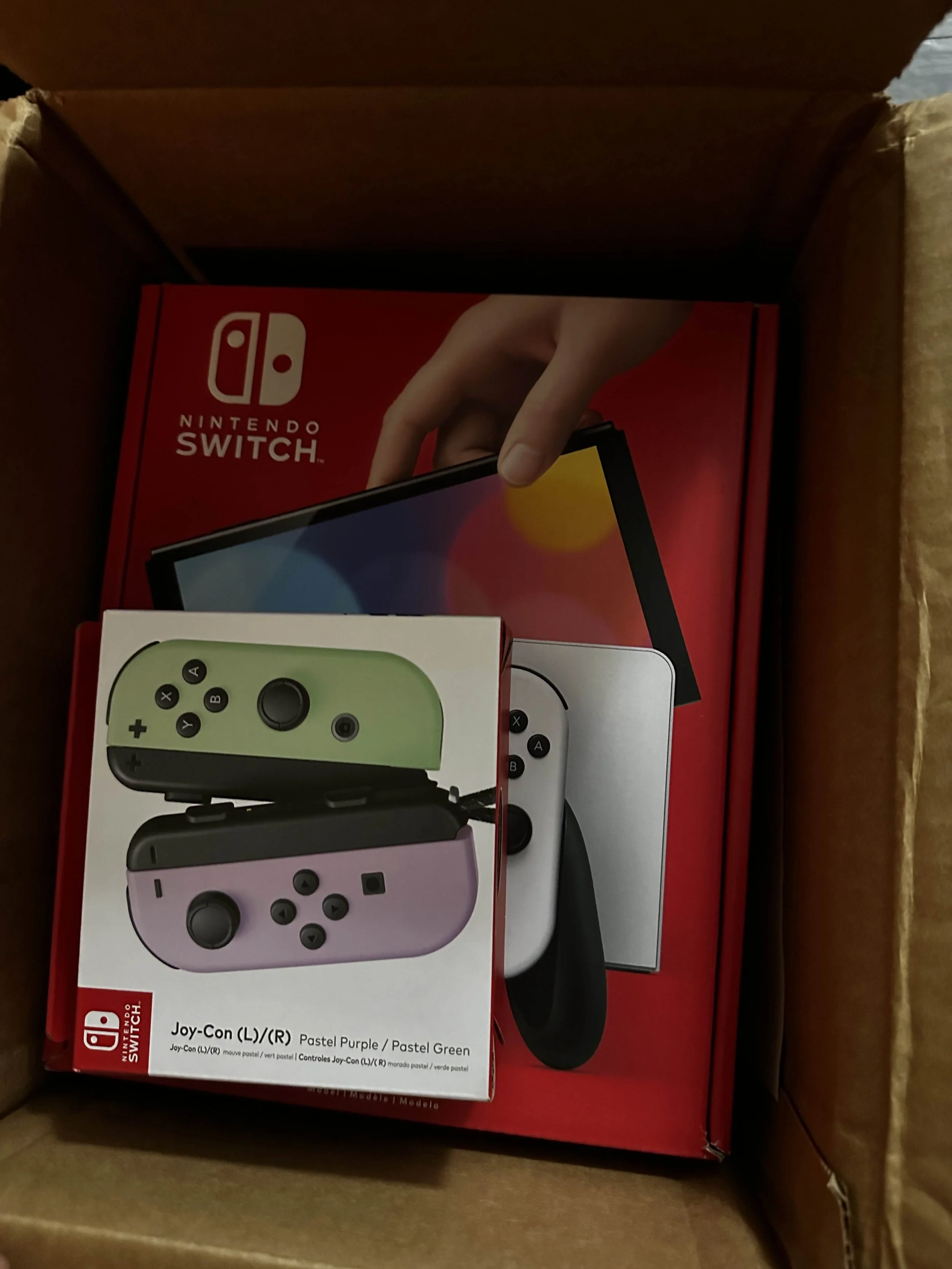 Open cardboard box containing a Nintendo Switch console, Joy-Con controllers in pastel purple and pastel green, and a box for the Nintendo Switch.