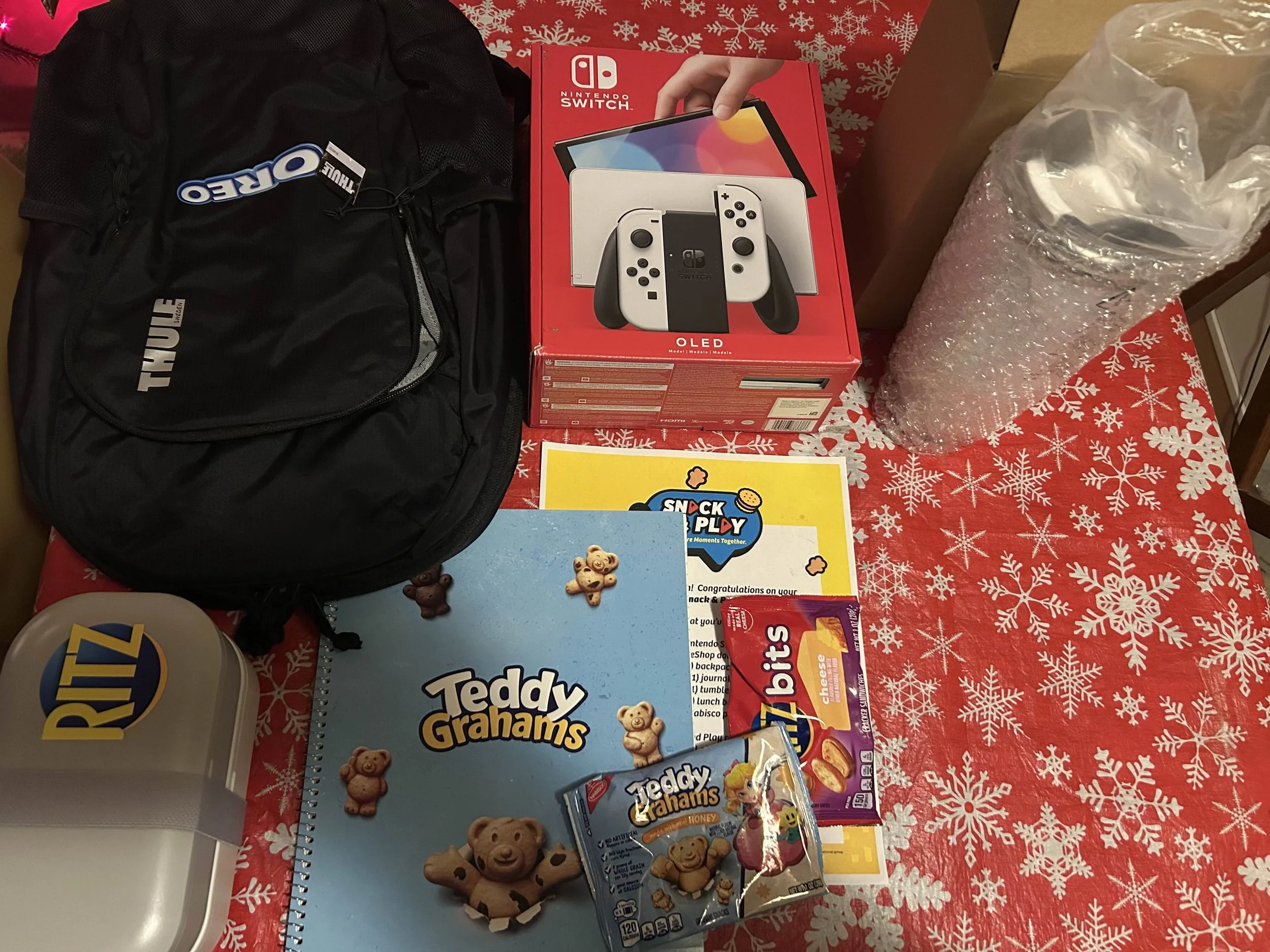 Collection of holiday gifts and items on a festive red tablecloth with white snowflakes, including a black Thule backpack, a Nintendo Switch OLED box, a large roll of bubble wrap, a container of Ritz Bits crackers, a Teddy Grahams notebook, and a pac