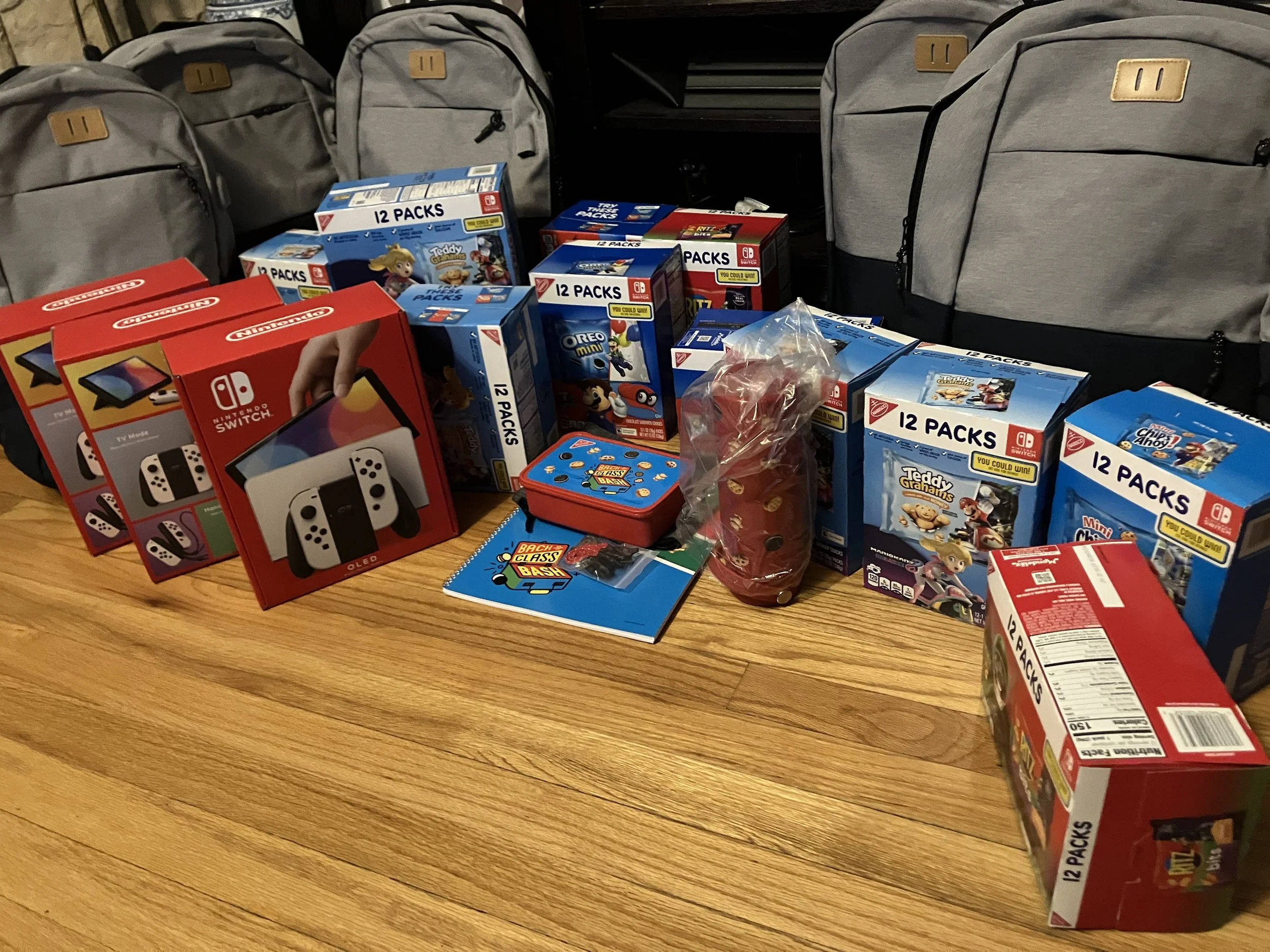 Table with multiple boxes of Nintendo Switch games, backpacks, and a small red and blue tin box. The games include titles like Teddygrams, Oreo, and Cheetos. The backpacks are grey with black zippers, arranged in the background.