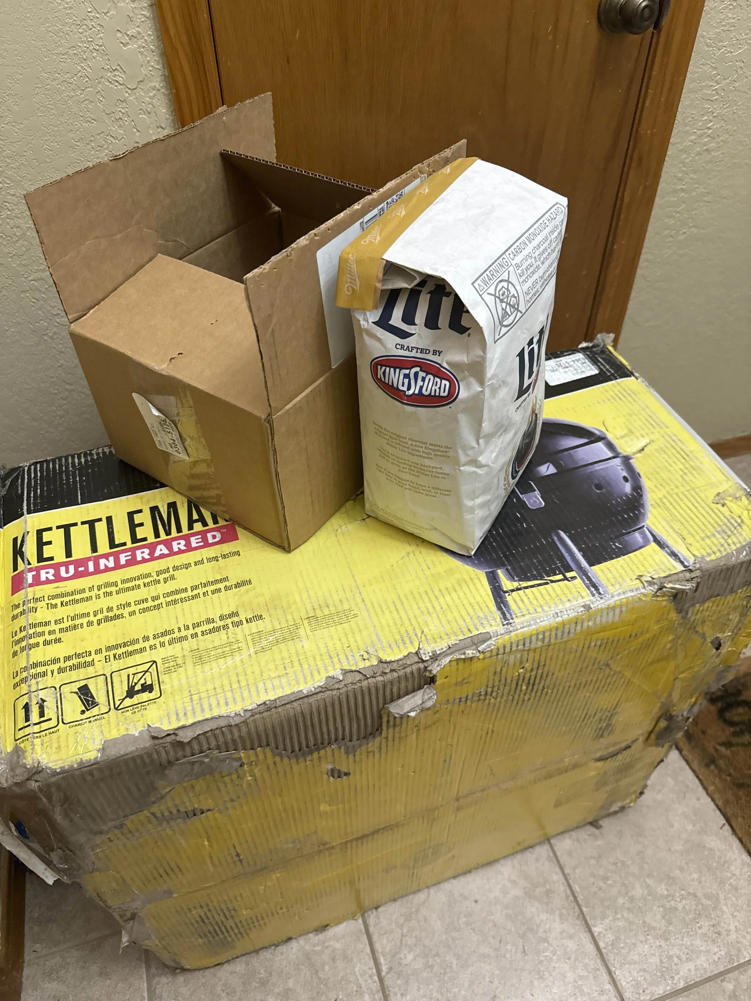 A partially opened cardboard box placed on a yellow Kettleman tri-infrared grill box with a bag of Miller Lite beer on top, against a wooden door and beige textured wall.