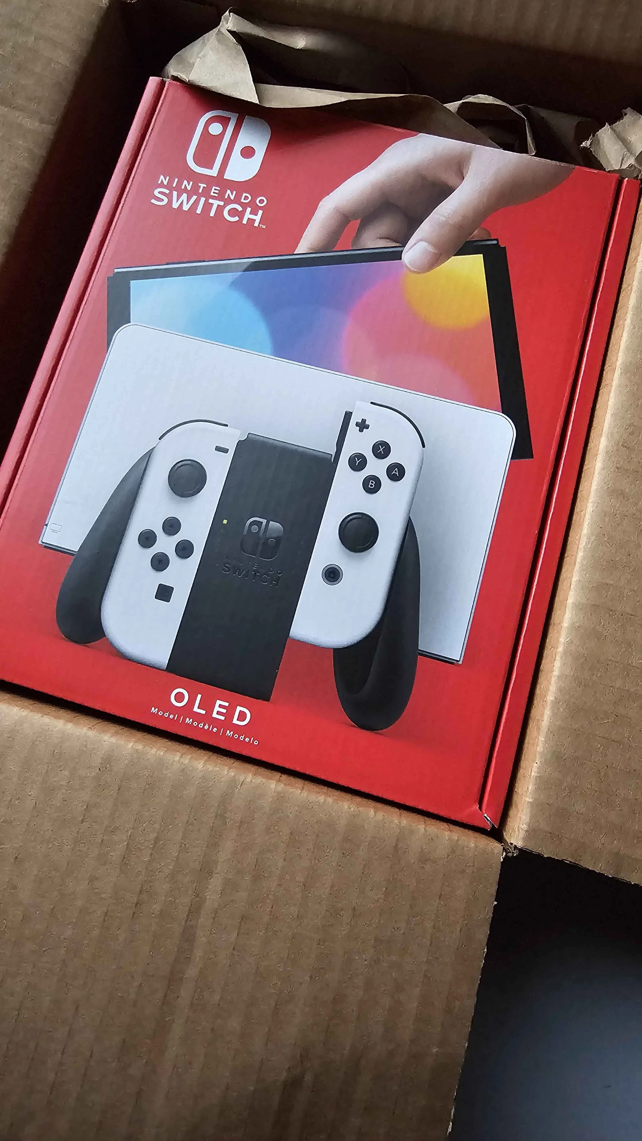 A boxed Nintendo Switch OLED gaming console with a white controllers and a black stand, inside a cardboard shipping container.