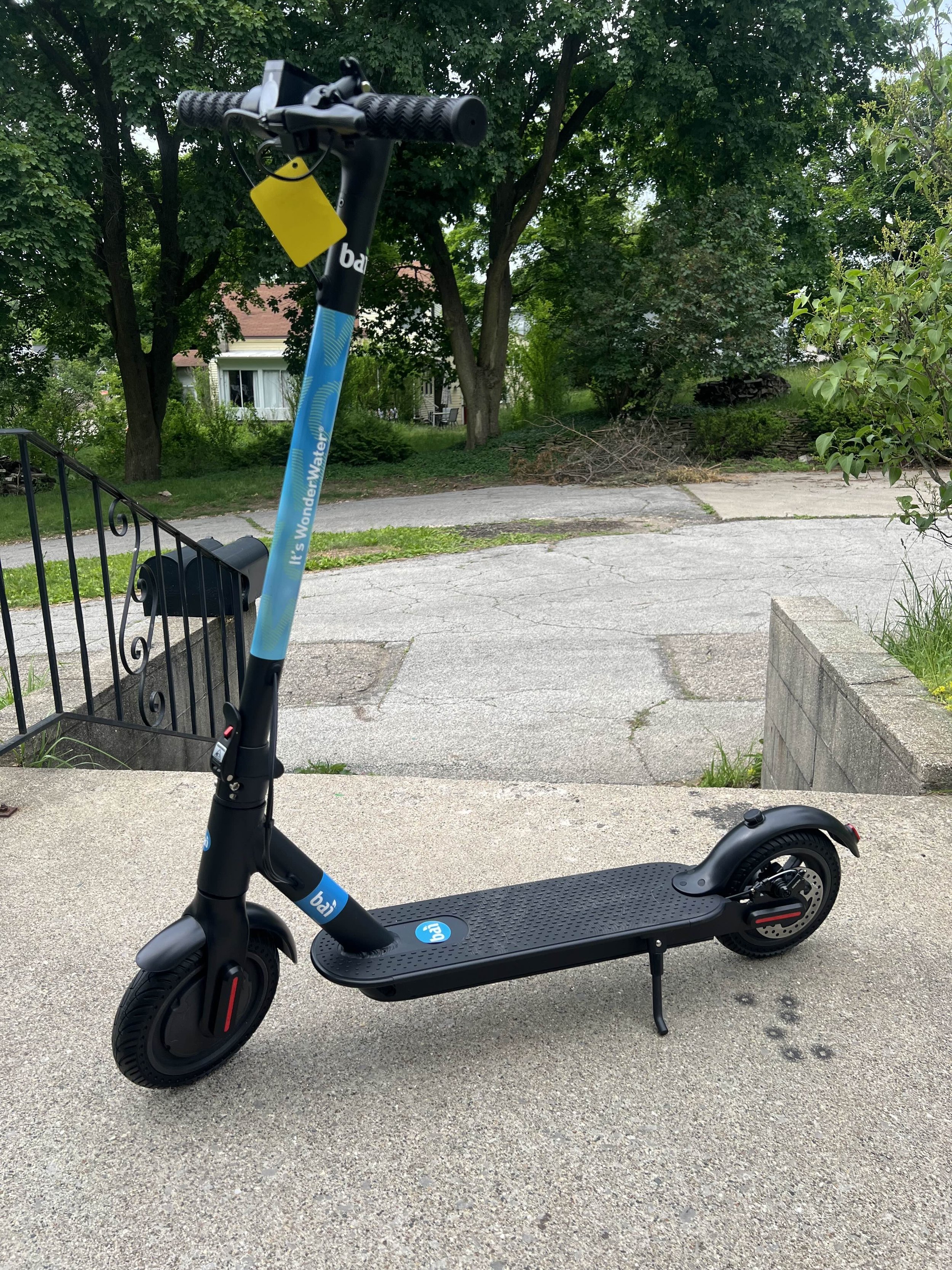 Black electric scooter parked on a concrete sidewalk with residential houses and trees in the background.