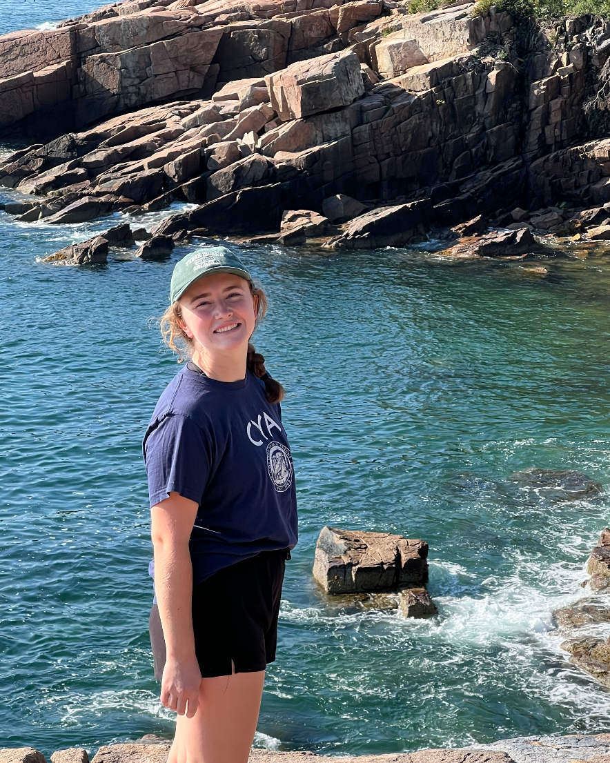 A young woman standing near rocky shoreline in front of ocean water, smiling at the camera, wearing a green cap, navy T-shirt, and black shorts.