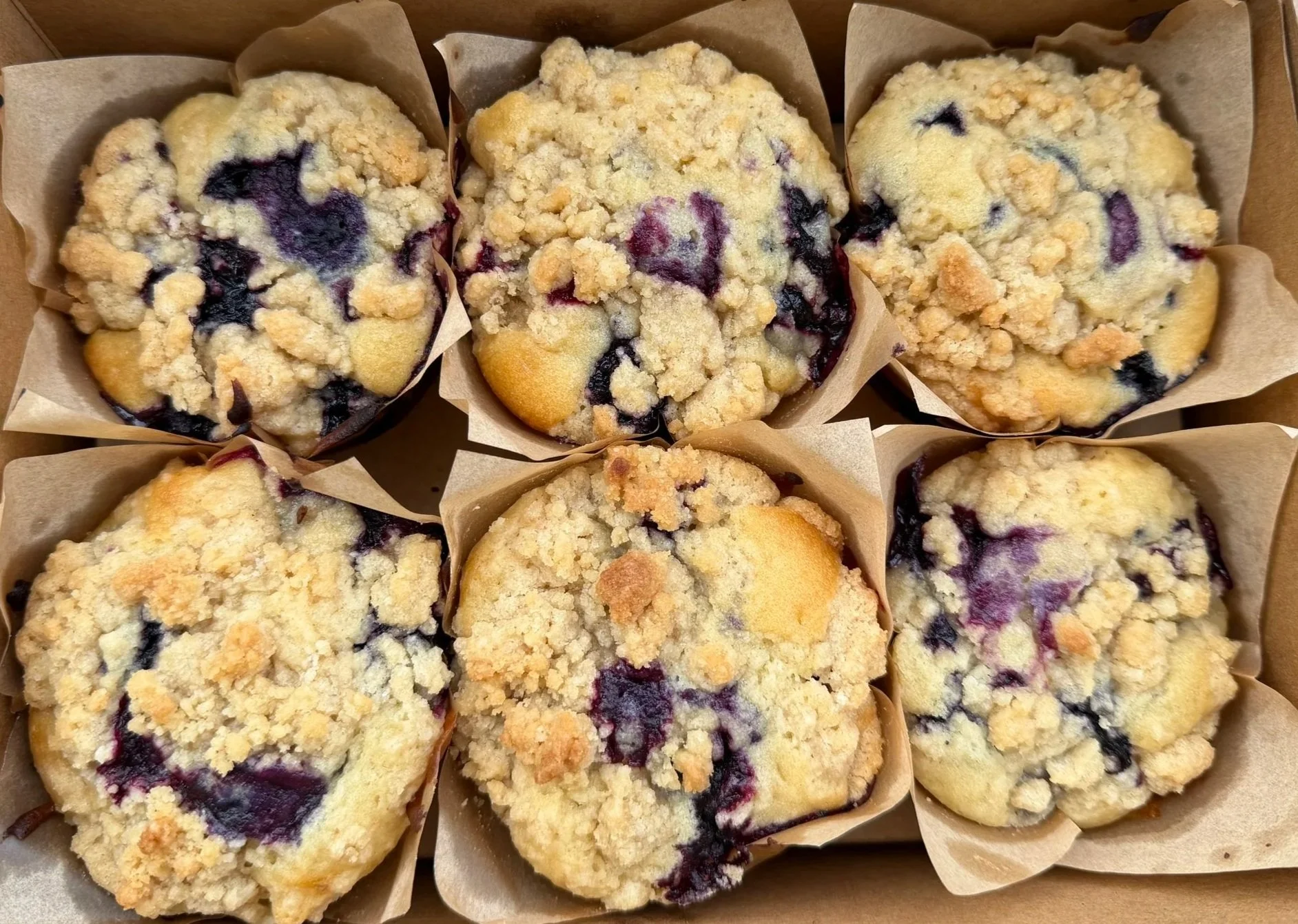Six blueberry muffins with crumb topping in parchment paper liners