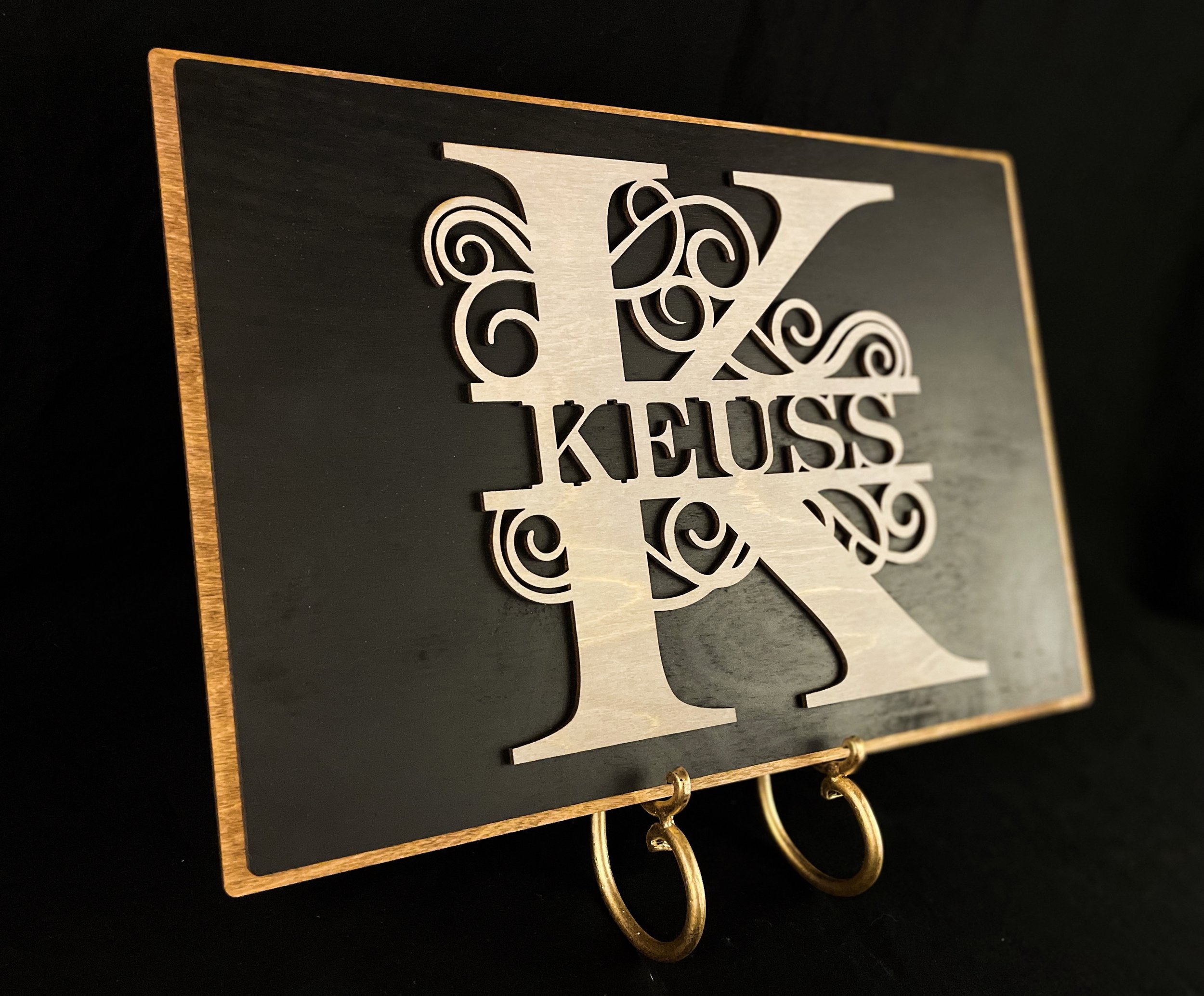 Decorative wall art with a large letter K and intricate scrollwork, featuring the name KEUSS in the center, mounted on a black background with a wooden border, and hanging hooks at the bottom.
