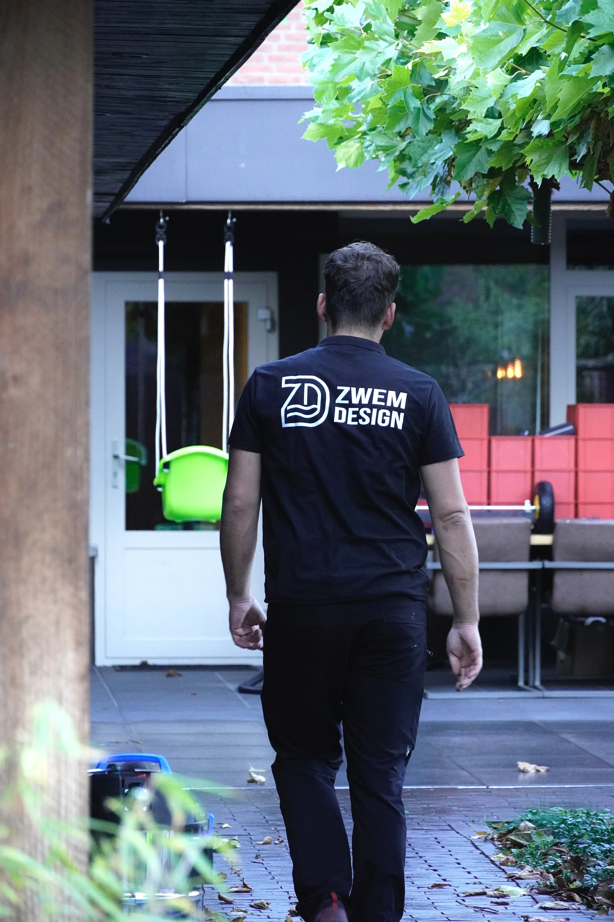 Man in zwart T-shirt met logo achter zich, wandelt op terras met tuinmeubelen en speelgoedkleuterstoel, buitenomgeving met planten en bomen.