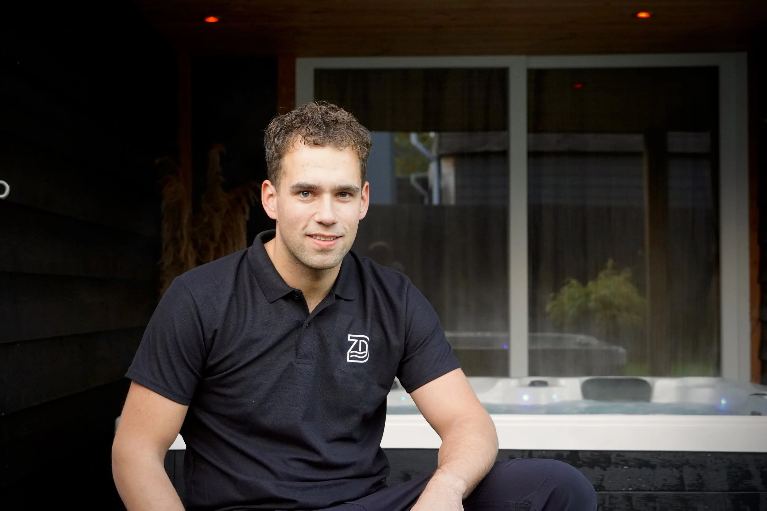een jonge man zit buiten bij een jacuzzi, draagt een zwart polo met een logo erop, en kijkt naar de camera