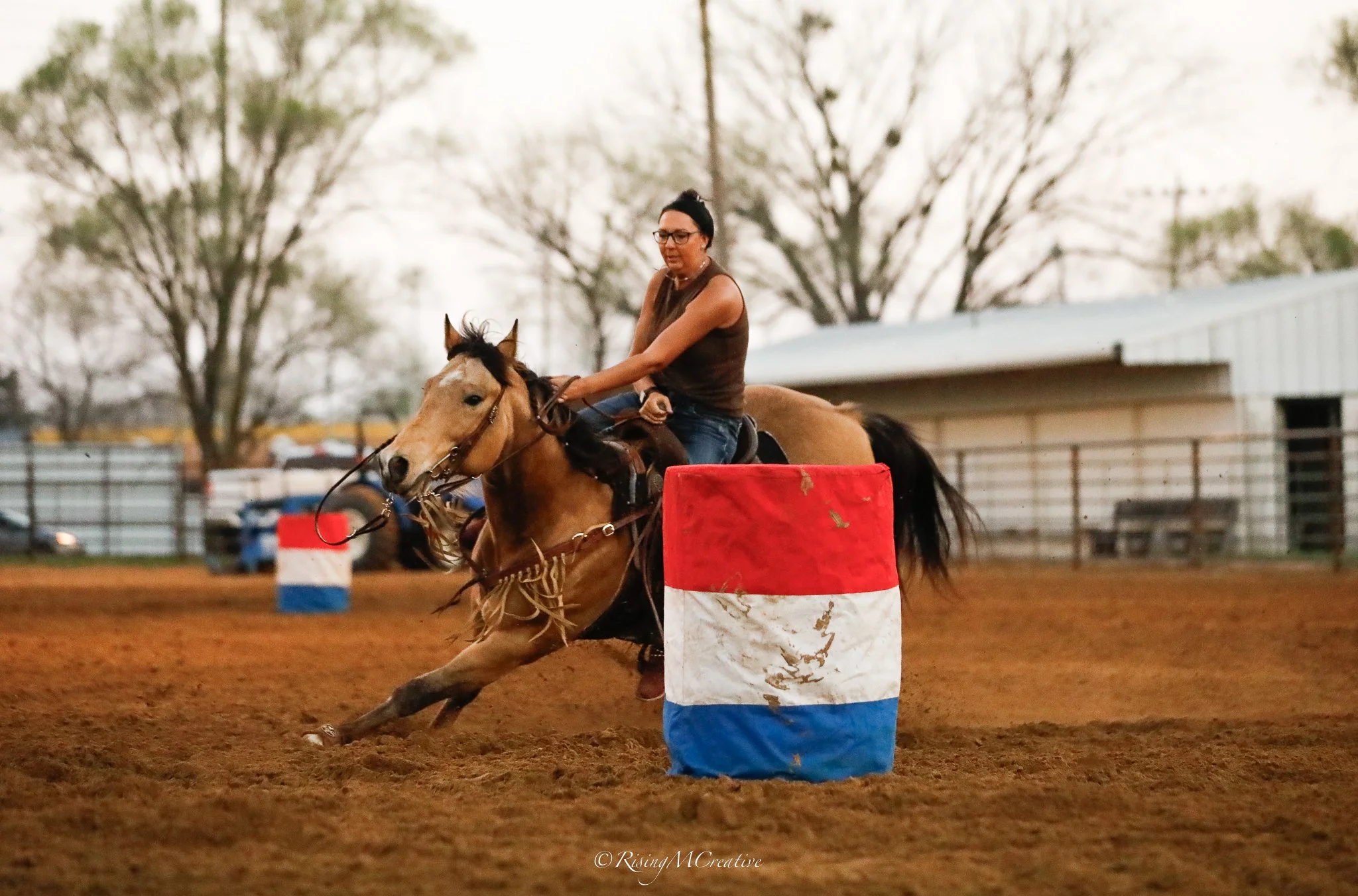 A woman riding a horse in a barrel racing competition, navigating around a red, white, and blue barrel on a dirt arena.