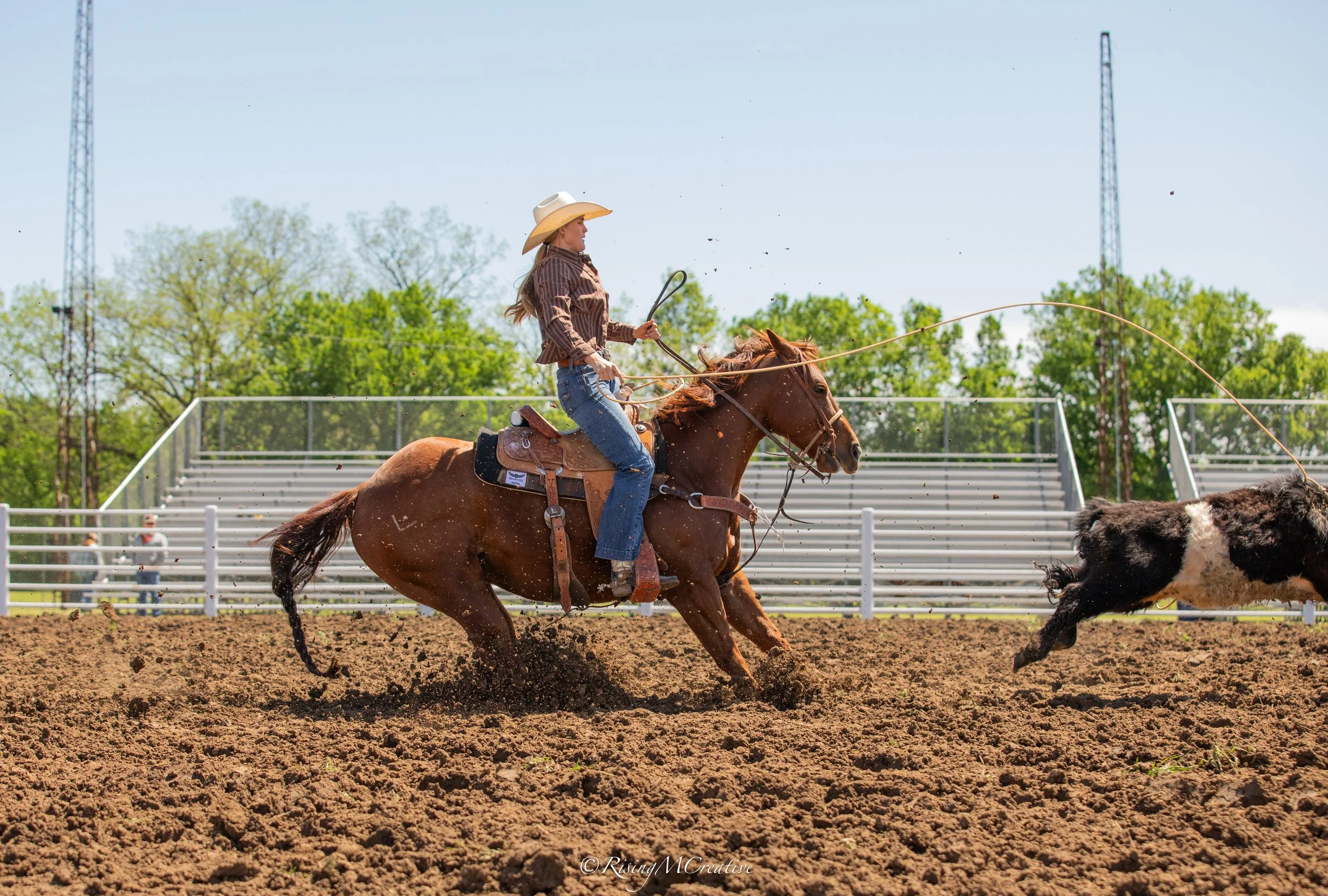 A woman wearing a cowboy hat and western attire rides a horse in a rodeo arena, her lasso swinging as she participates in a rodeo event, with onlookers and empty bleachers visible in the background.