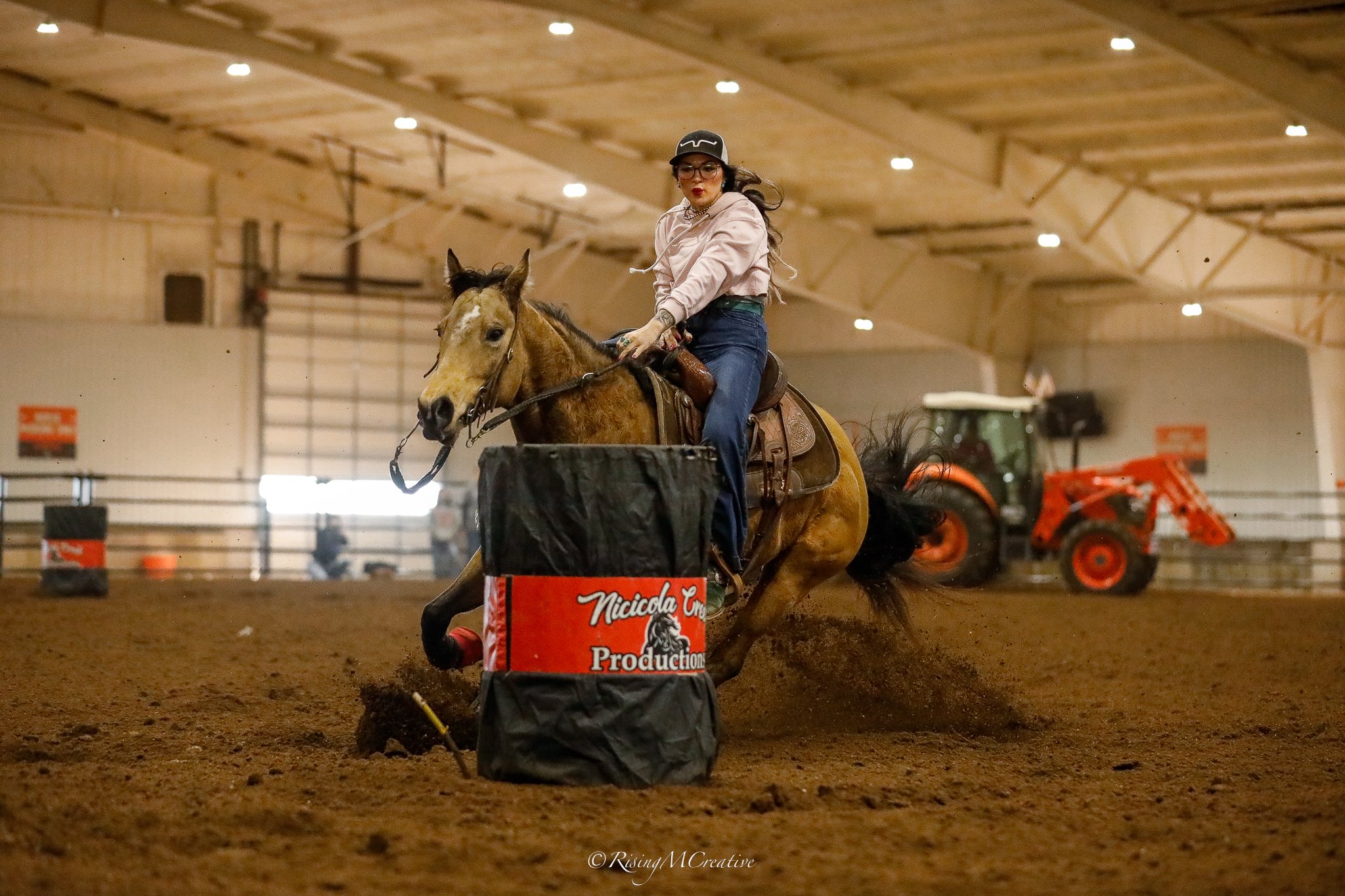 A woman riding a horse in an indoor riding arena, jumping over a barrel obstacle with the words 'Nicolina Cross Production' on it. She is wearing glasses, a cap, and casual riding attire.