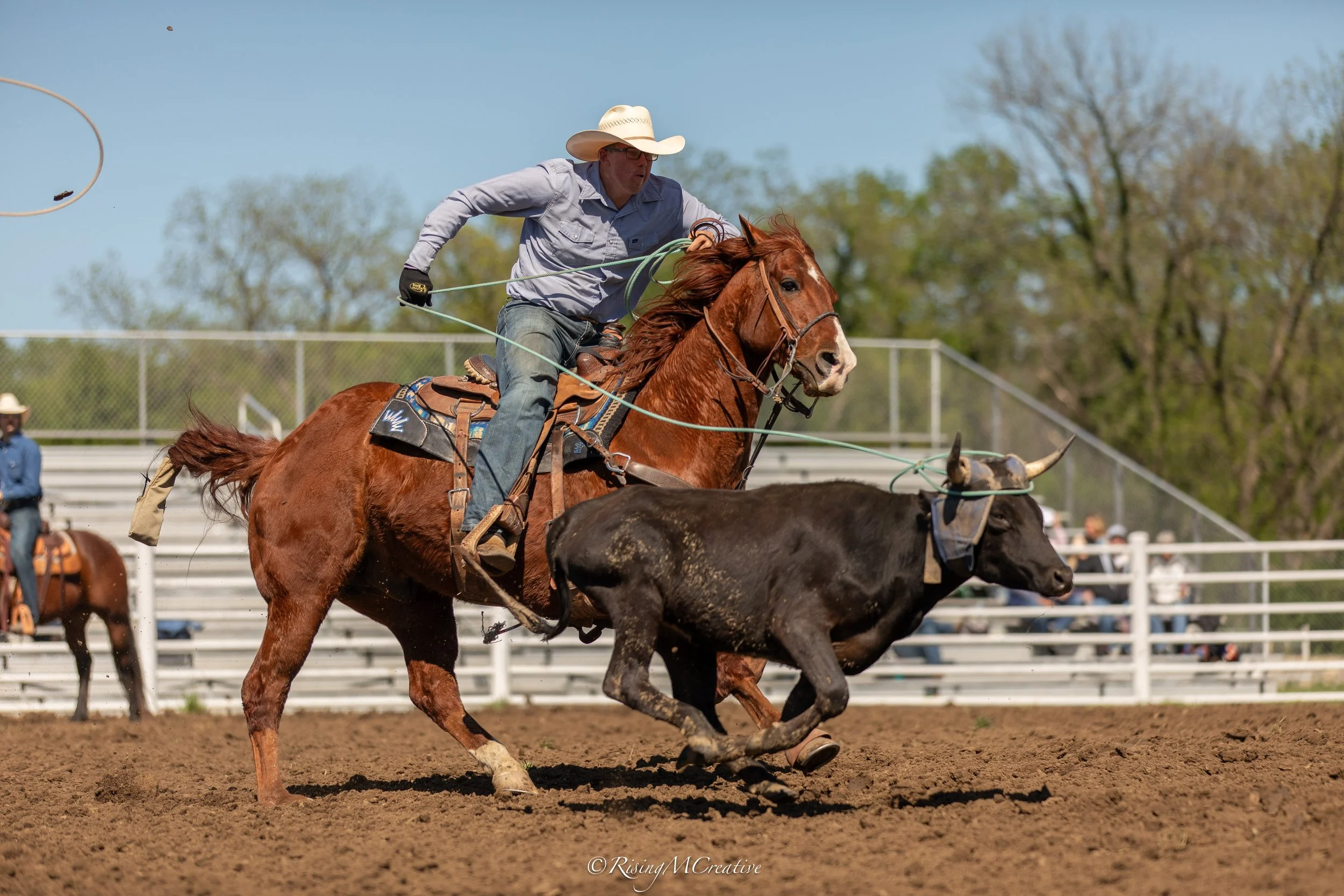 A cowboy wearing a cowboy hat, glasses, and a light gray shirt, riding a galloping horse during a rodeo or horse race, with a fencing and trees in the background on a sunny day.