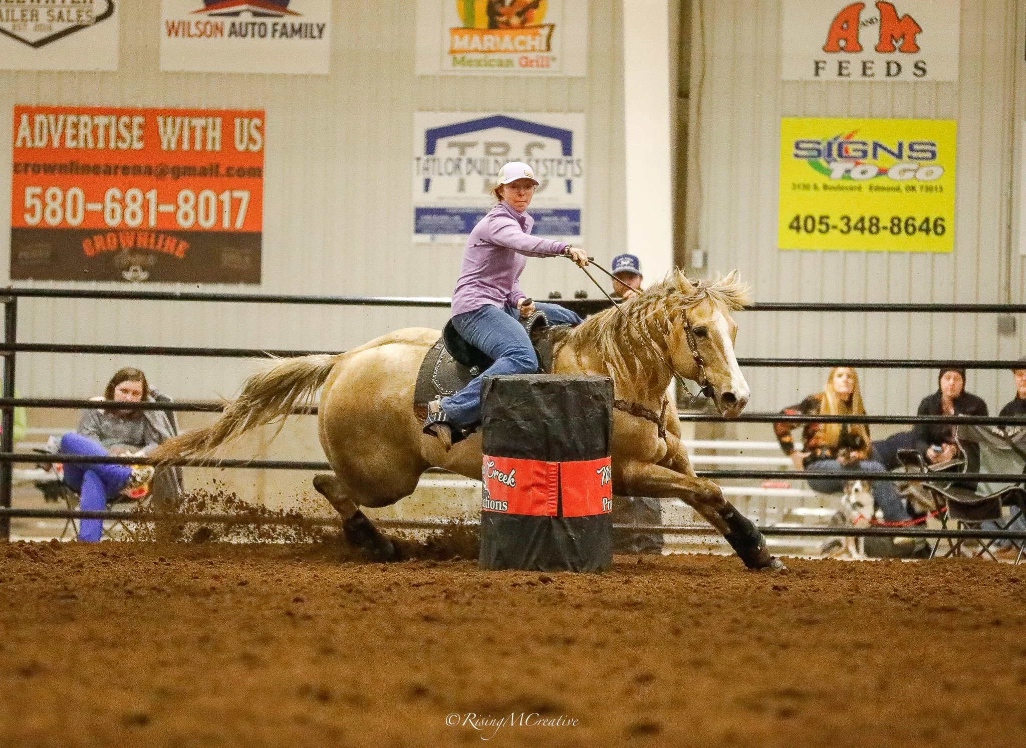 A person riding a bucking horse in an indoor arena, with advertisements and spectators in the background.