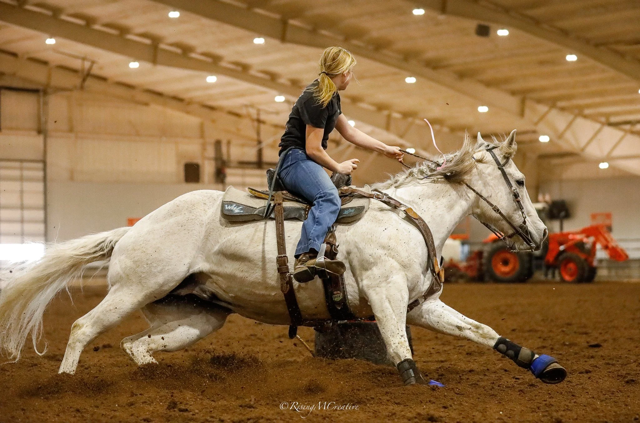 A person riding a galloping palomino horse in an indoor arena. The rider is wearing a purple sweatshirt, jeans, and a white baseball cap, and is holding a lasso while leaning forward as the horse runs around a barrel marked "Pine Creek Productions."