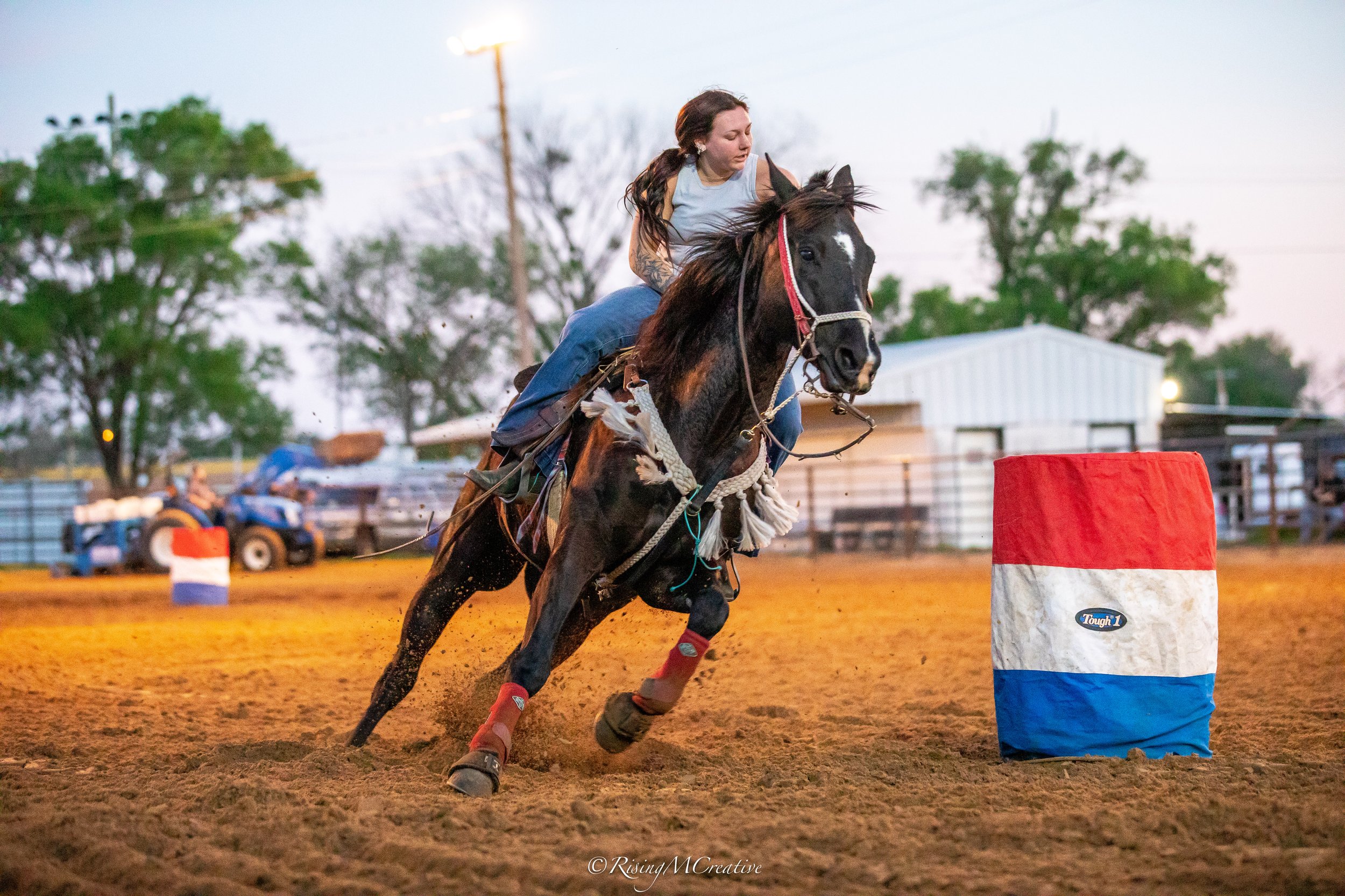 A female horseback rider navigating a barrel racing course on a black horse at dusk, with a barrel in the foreground and a fenced arena in the background.