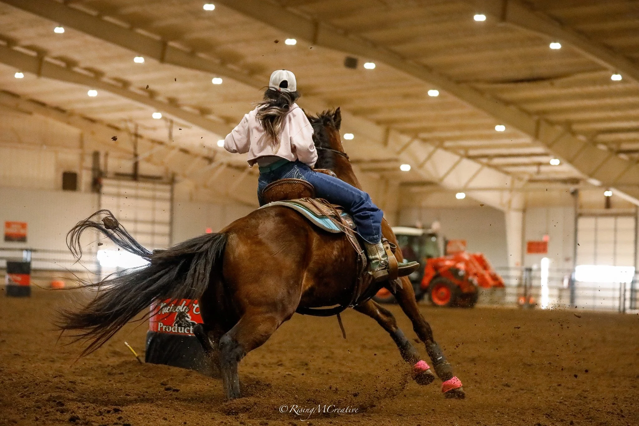 A woman wearing a white cap and pink jacket rides a brown horse racing inside an indoor arena with a dirt floor.