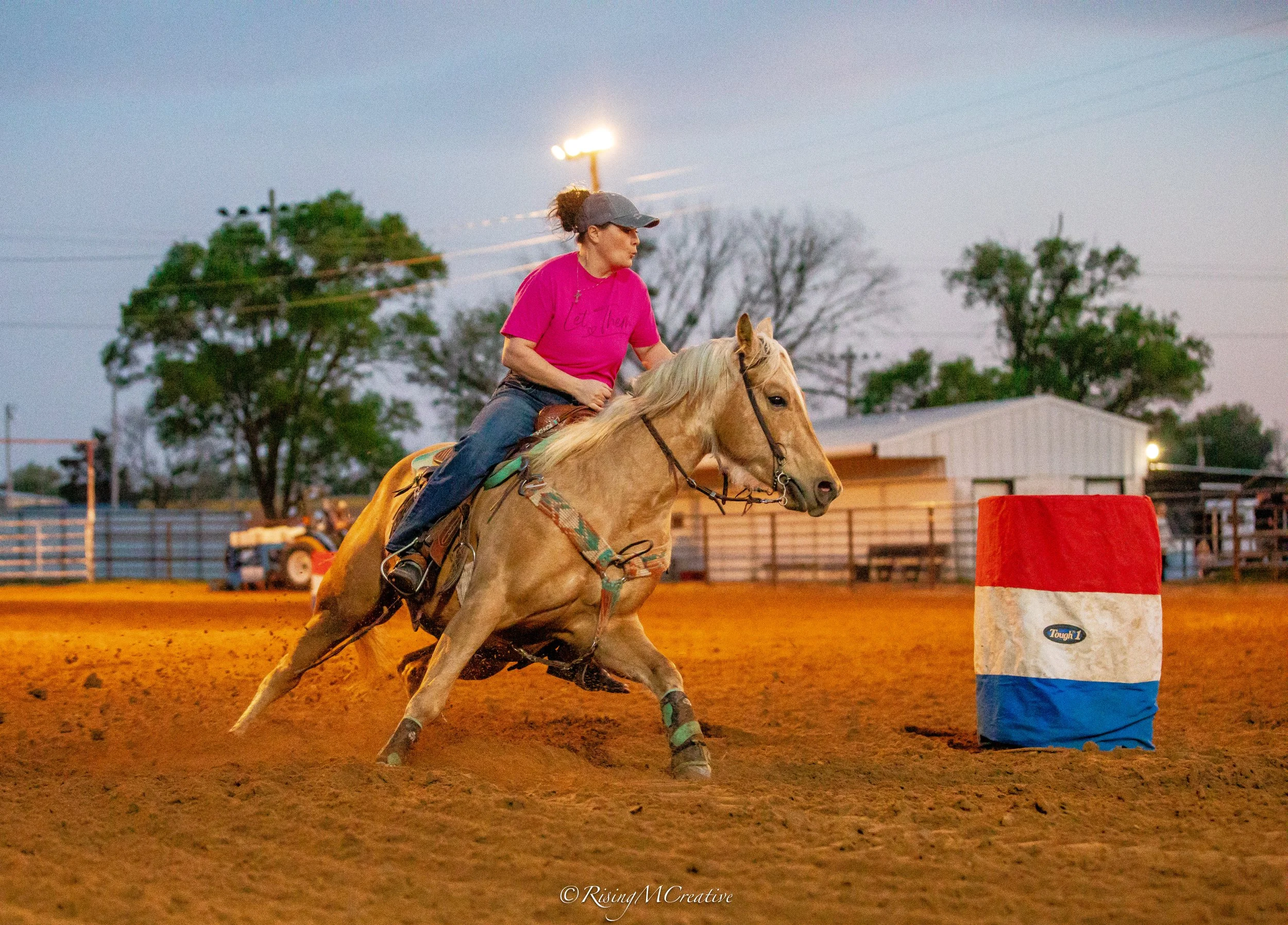 A woman wearing a pink shirt and gray cap rides a galloping horse during a rodeo event at dusk, navigating around a red, white, and blue barrel obstacle.