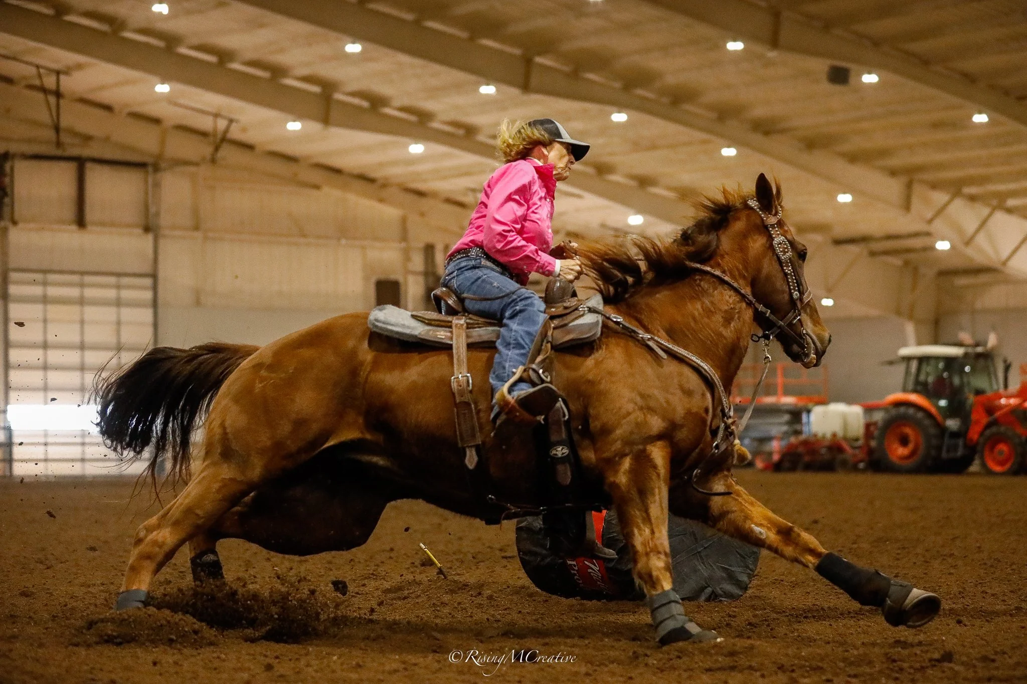 A woman in a pink jacket and jeans rides a bucking bronco horse in an indoor arena, with a low ceiling and bright overhead lights.