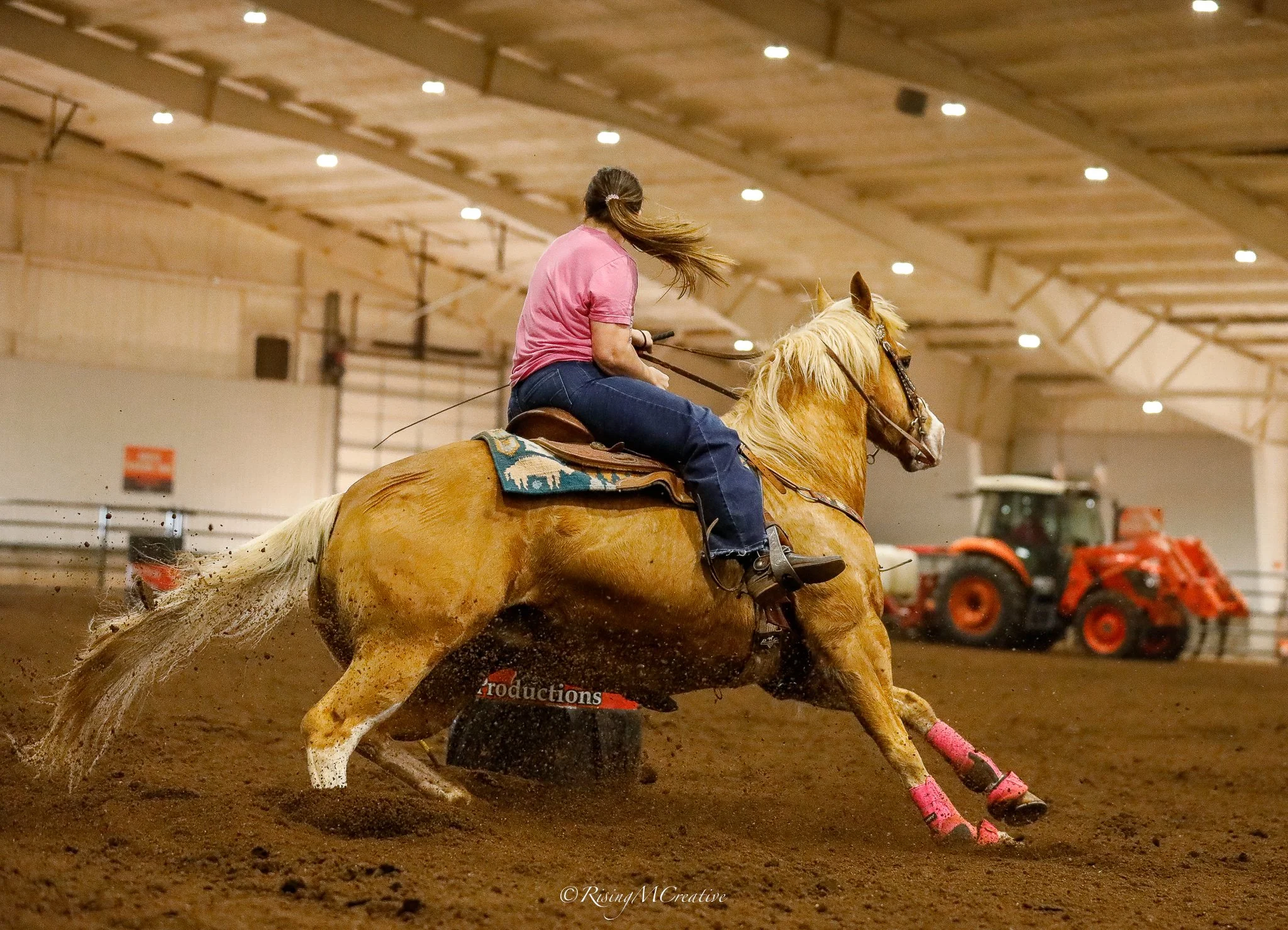 A woman riding a galloping palomino horse inside an indoor arena, with Tractor equipment in the background.