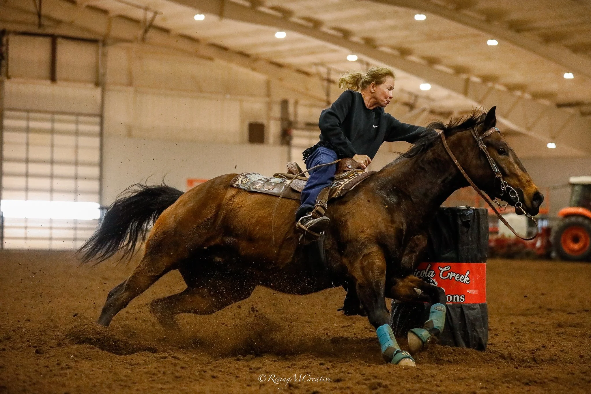 A female equestrian rider in black jacket and jeans riding a brown horse with black mane inside an indoor arena, featuring dirt floor, bright ceiling lights, and equipment in the background.