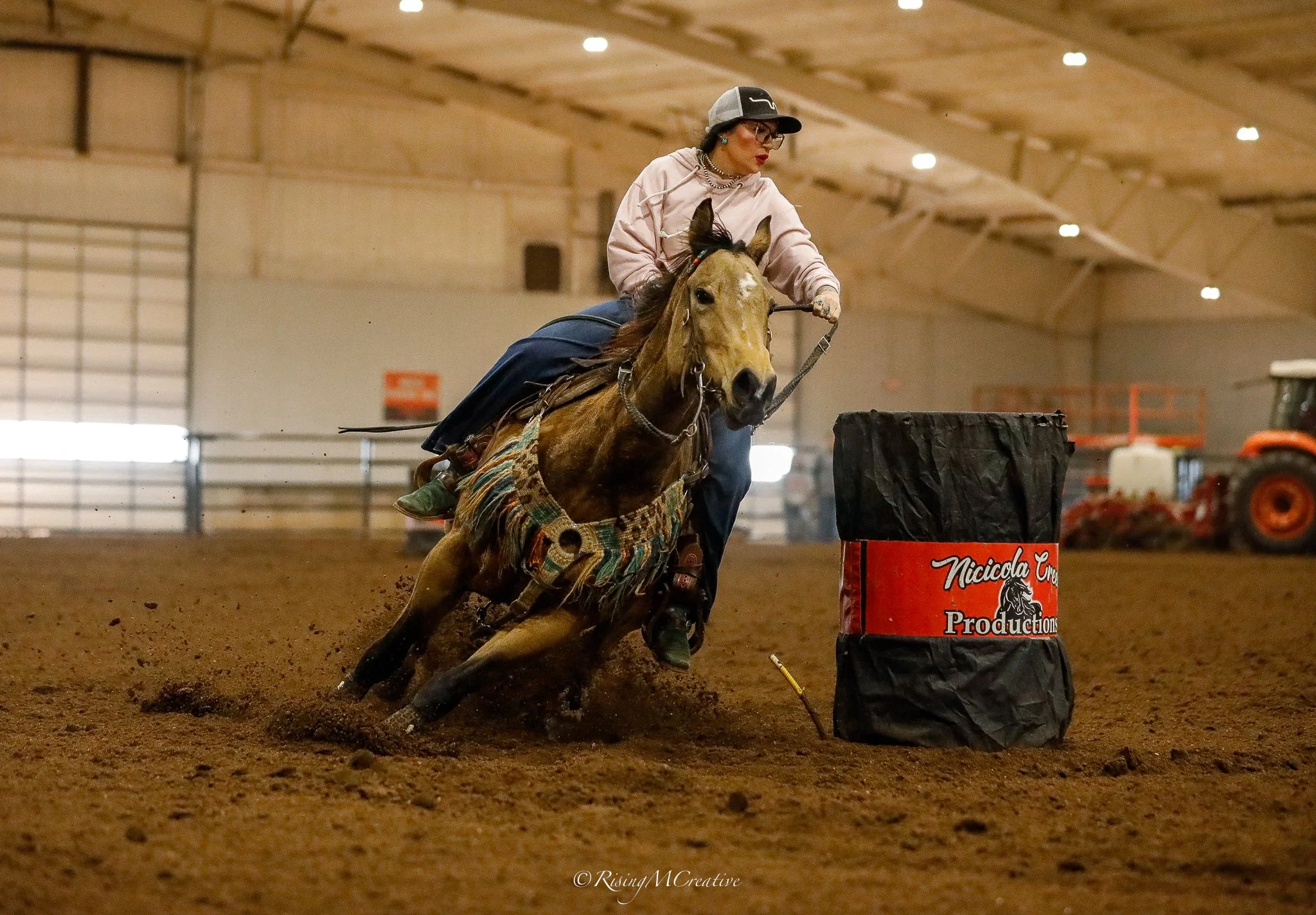 A woman riding a horse inside an indoor arena, navigating around a black barrel marked 'Nicola Creations.' The horse is kicking up dirt as they go around the barrel.