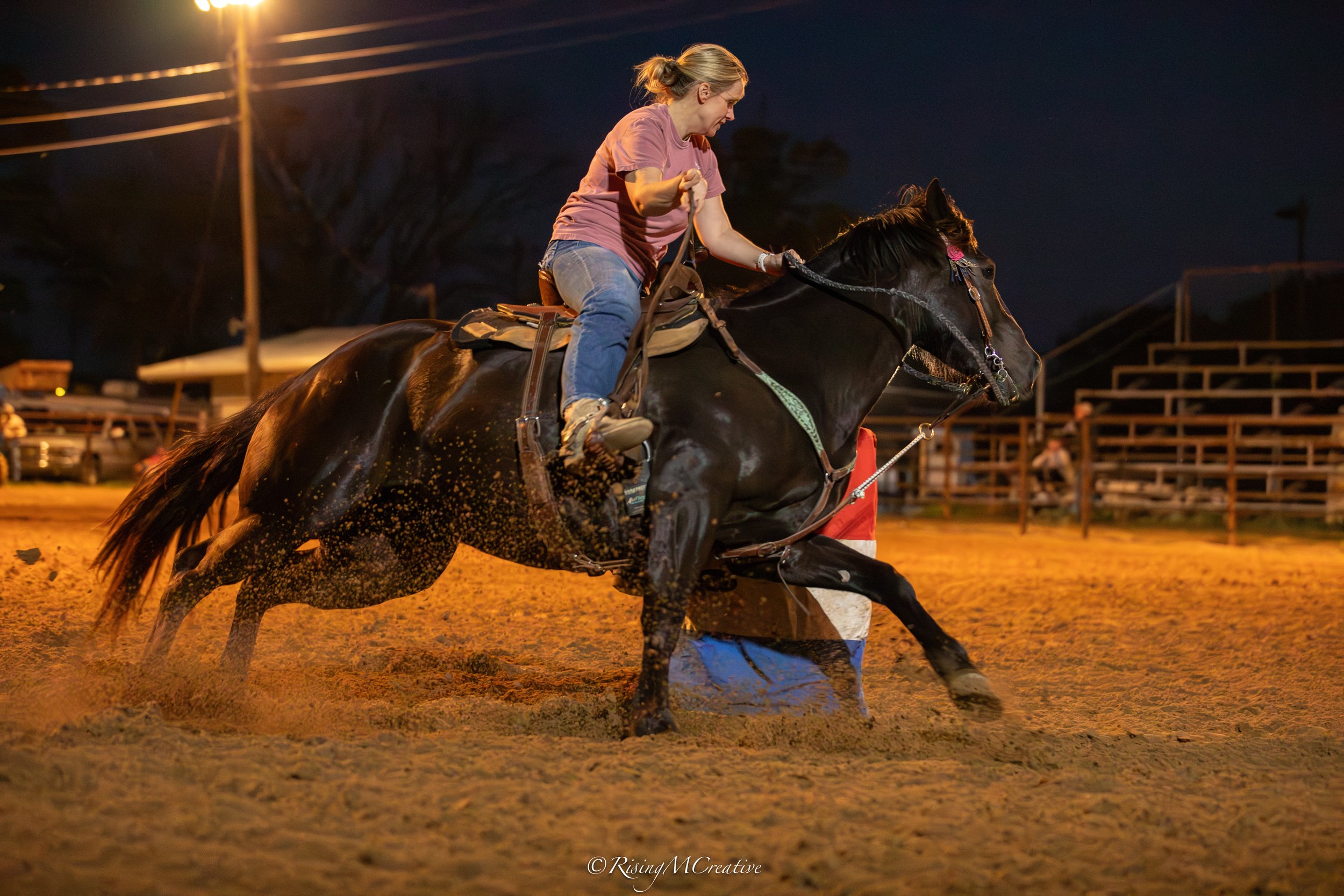 A woman riding a galloping horse at night in an outdoor arena.