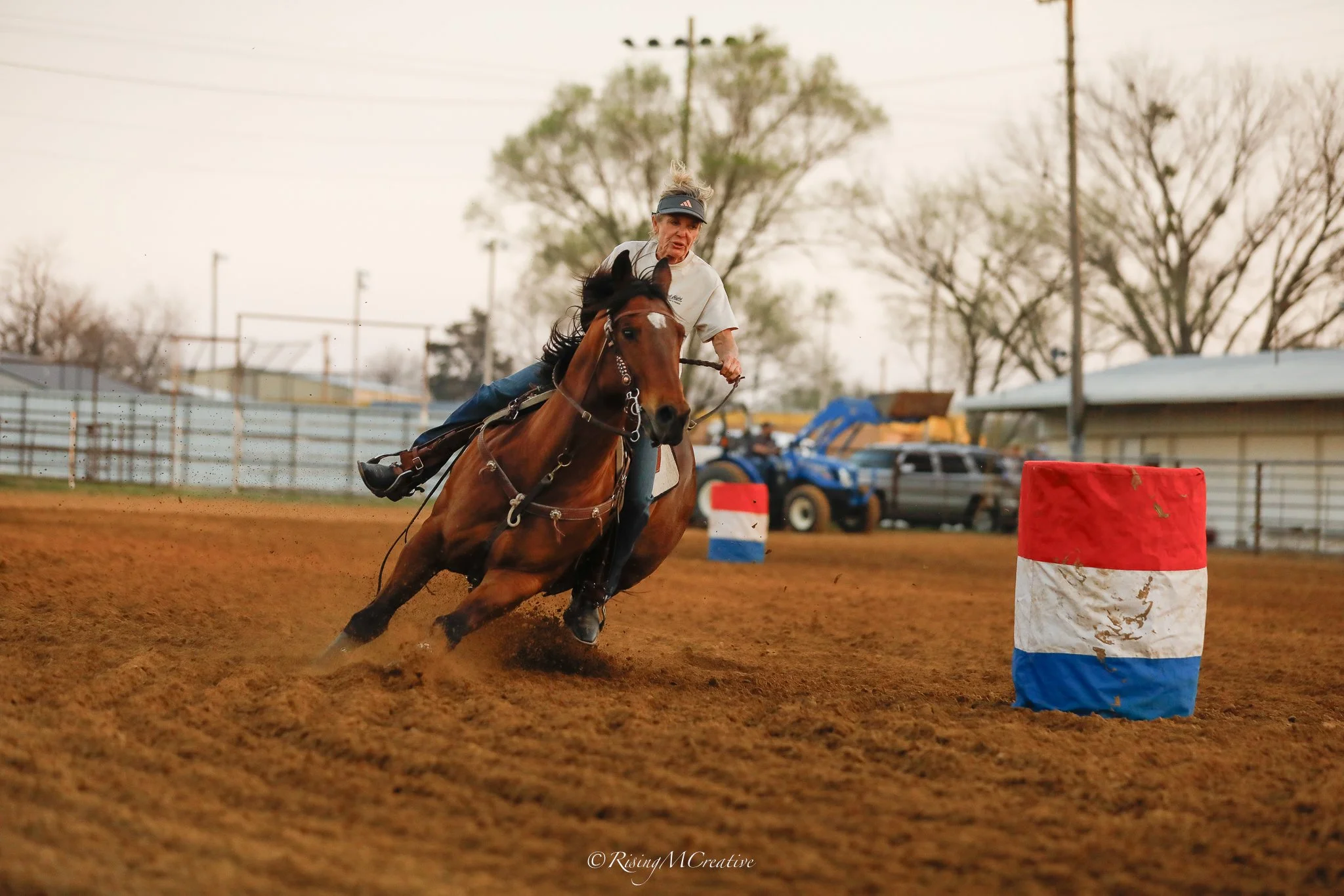 An elderly woman wearing a visor hat and white shirt rides a brown horse in a dirt arena, navigating around orange and white barrels during a barrel racing event.