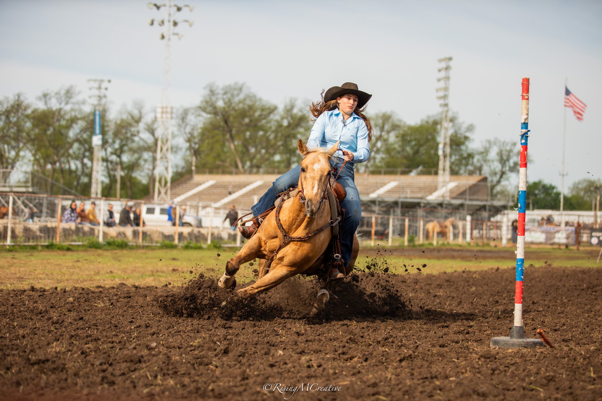 A woman riding a galloping horse on a dirt track, navigating a pole during a barrel racing event at a rodeo arena. She is wearing a black hat and a light blue shirt.