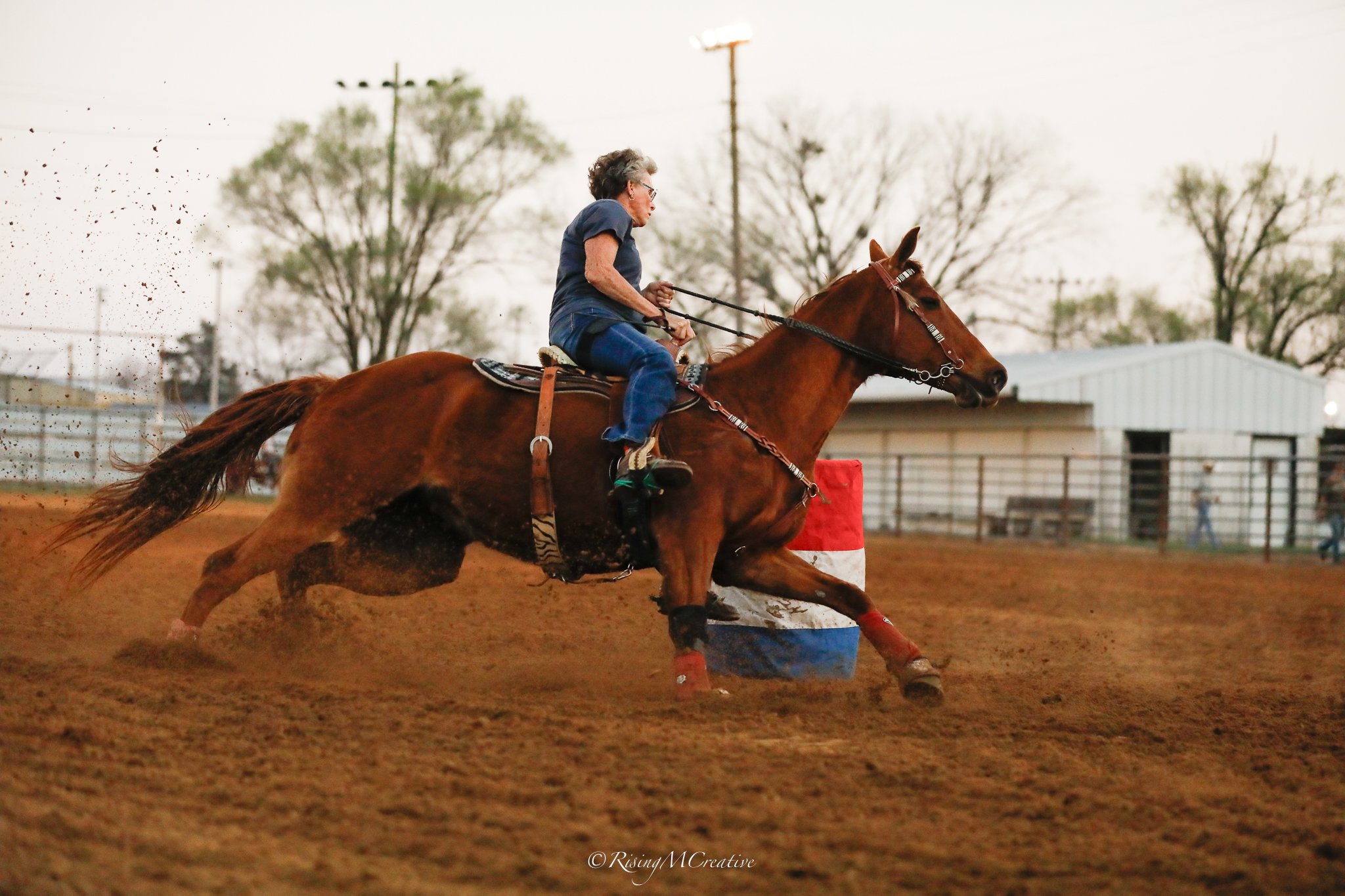 An elderly woman riding a galloping brown horse during a barrel racing competition on an outdoor dirt arena.