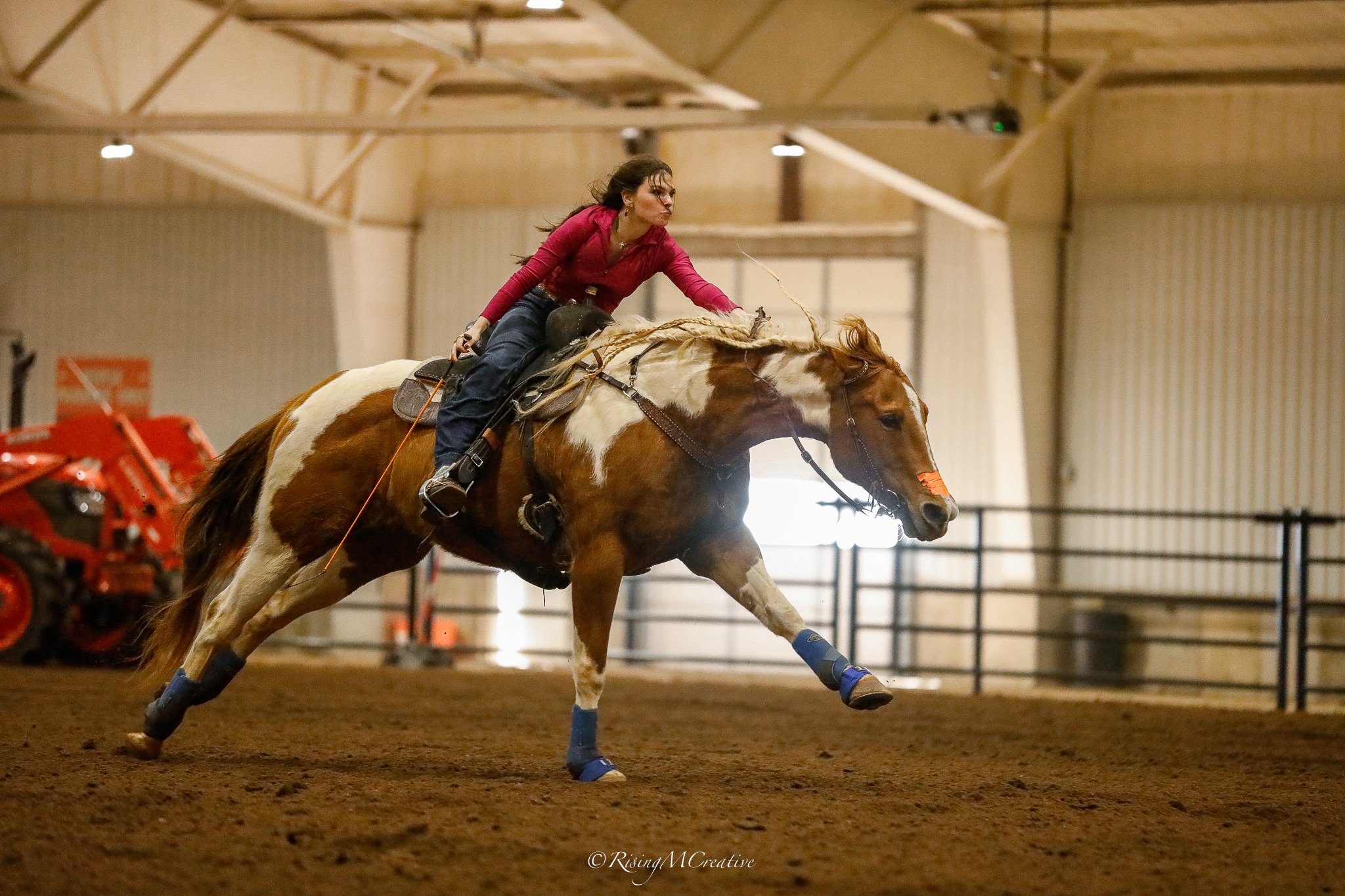 A woman riding a brown and white horse inside an indoor arena, participating in a rodeo event. The horse is jumping over a blue obstacle.