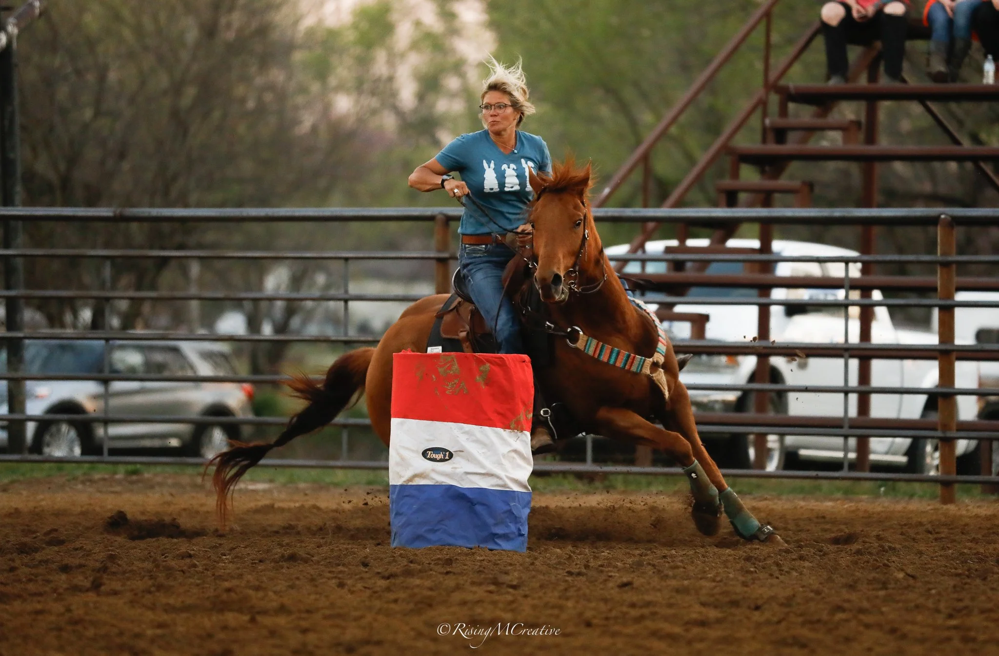 A woman riding a horse in an indoor arena, wearing glasses and a blue shirt, as she navigates around a barrel, with a wooden arena stands and trees in the background.