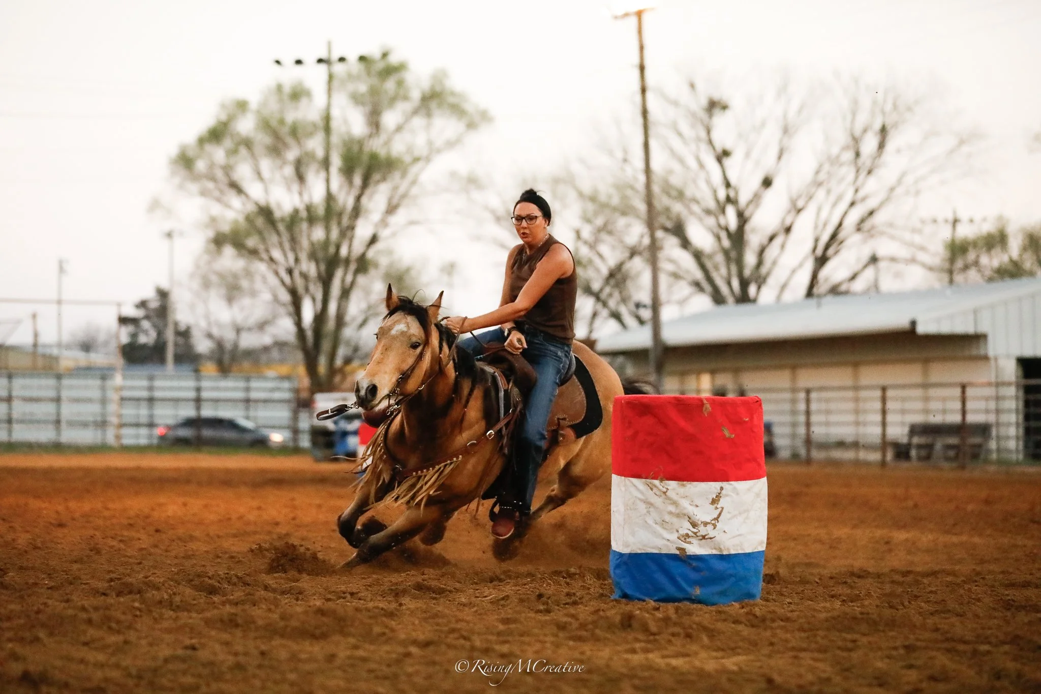 A woman riding a horse around a barrel during an equestrian event at an outdoor arena, with trees and a fence in the background.