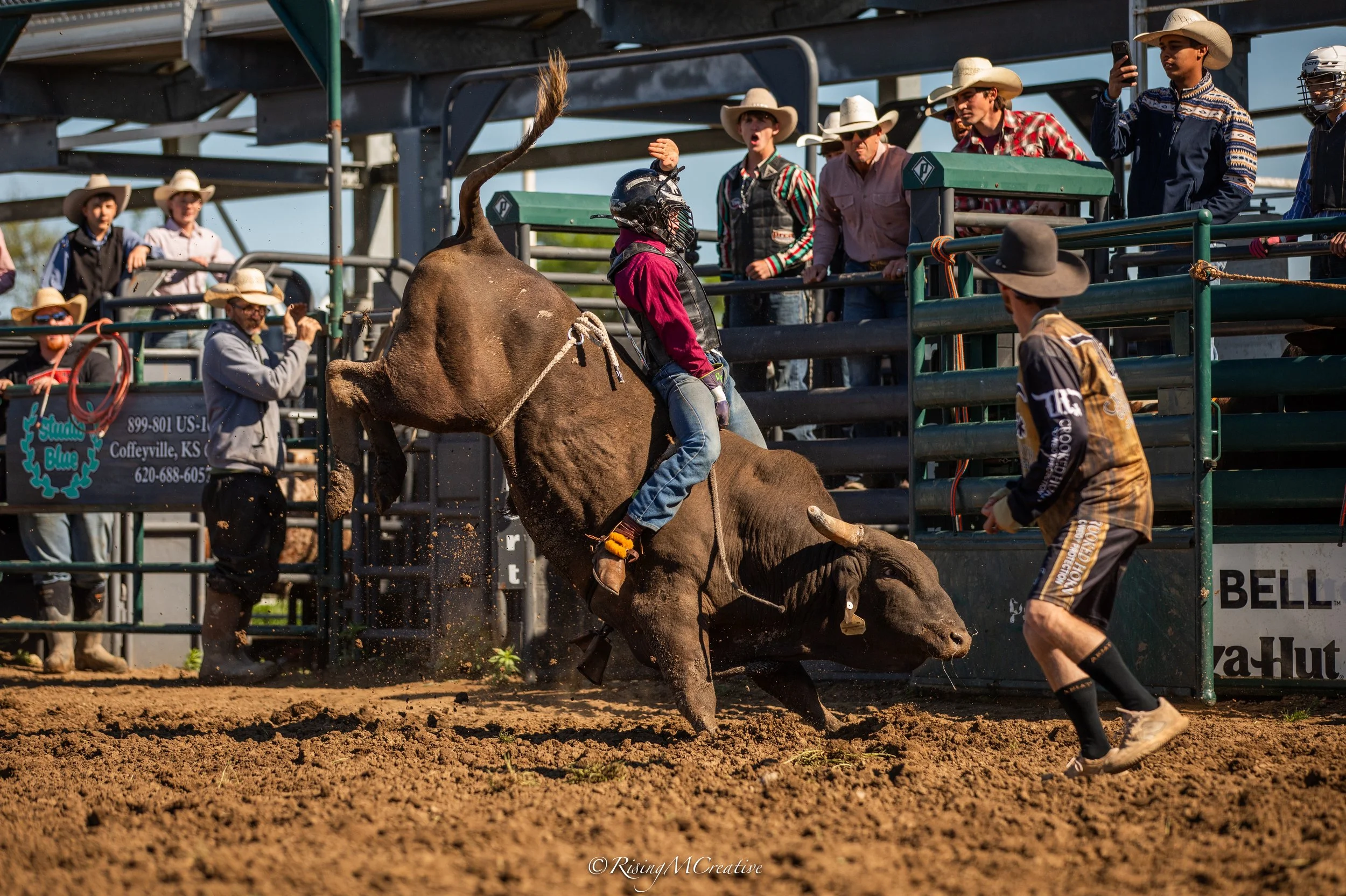 A rodeo scene where a bull rider wearing a helmet and pink shirt rides a bucking bull, with a man in cowboy attire holding a rope nearby. Several spectators, mostly wearing cowboy hats, observe from the stands and surrounding area. The event takes pl