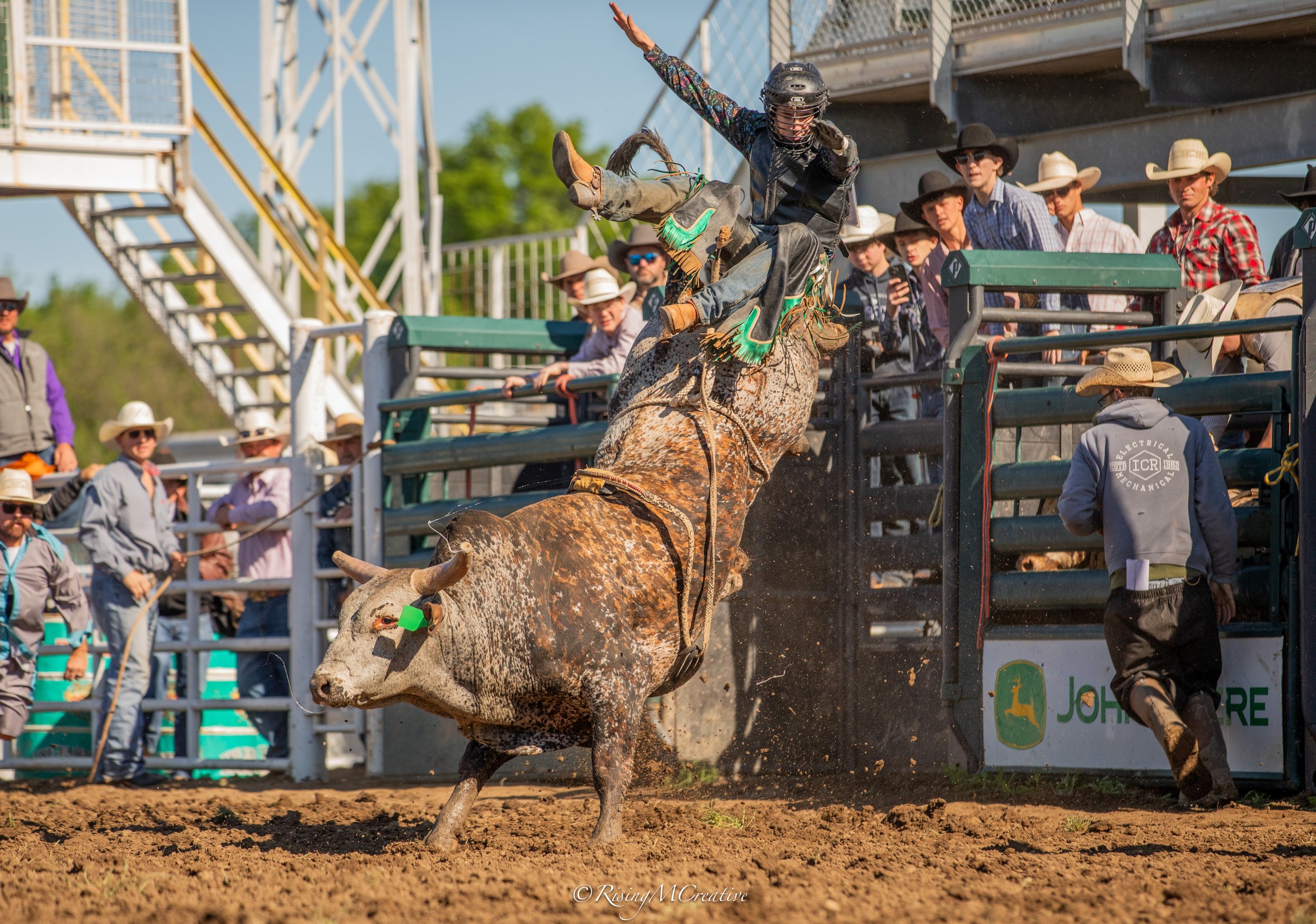 A rodeo cowboy riding a bucking bull with a crowd watching spectators, some wearing cowboy hats, behind the rodeo fence. The cowboy is wearing a helmet and western attire. The event takes place outdoors on dirt ground.