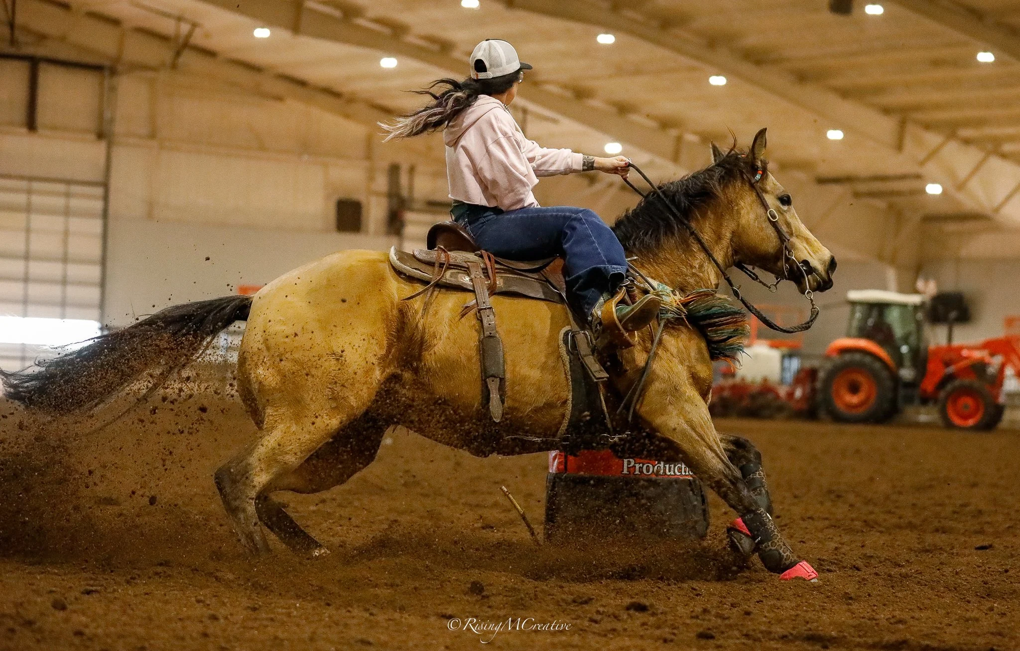 A woman wearing a baseball cap, hoodie, and jeans riding a tan horse inside an indoor arena. The horse is kicking up dirt as it runs around a barrel, likely participating in a barrel racing event. There are bright ceiling lights and orange equipment 