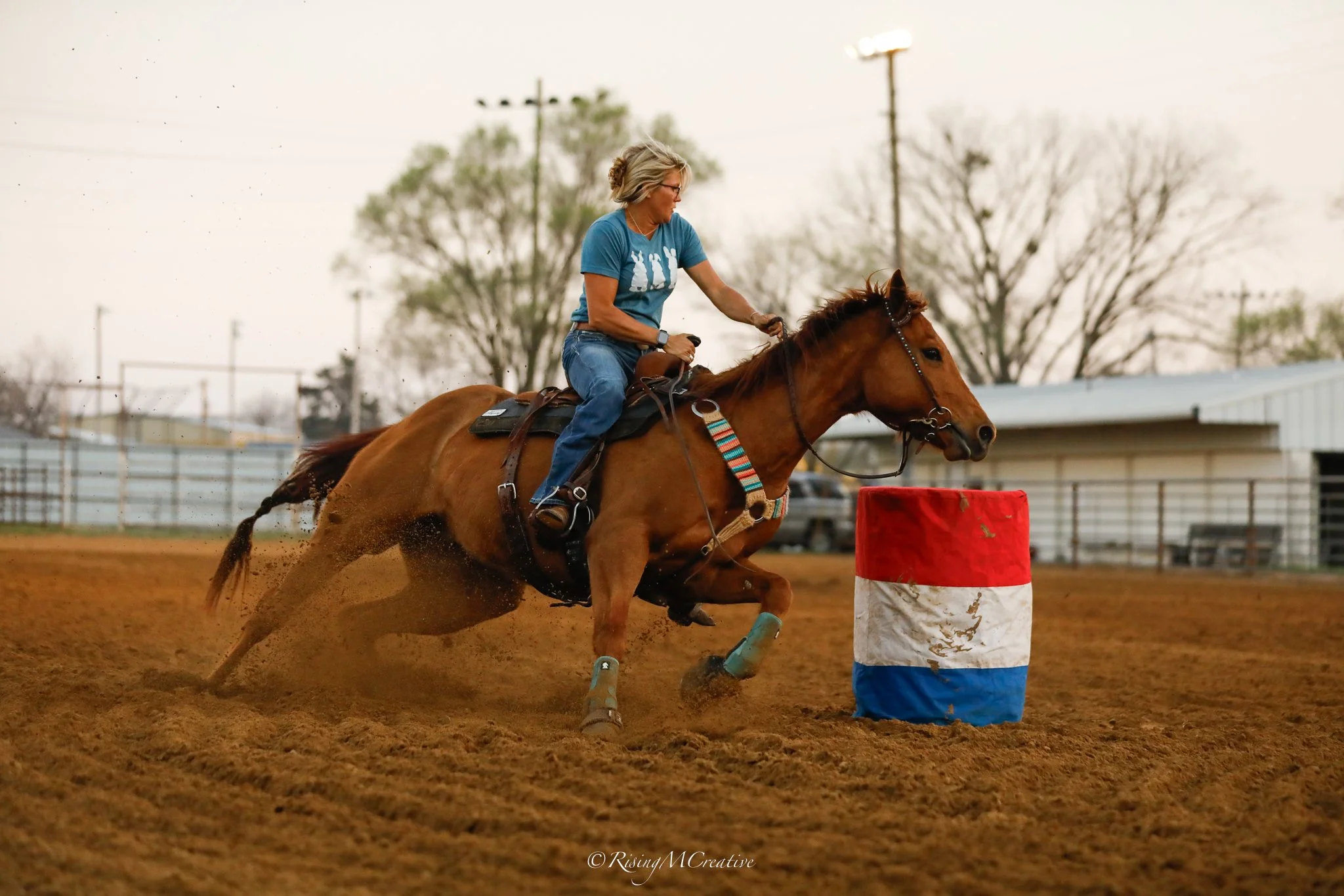 A woman wearing a blue t-shirt riding a galloping horse around a red and white barrel during an equestrian event on dirt ground.