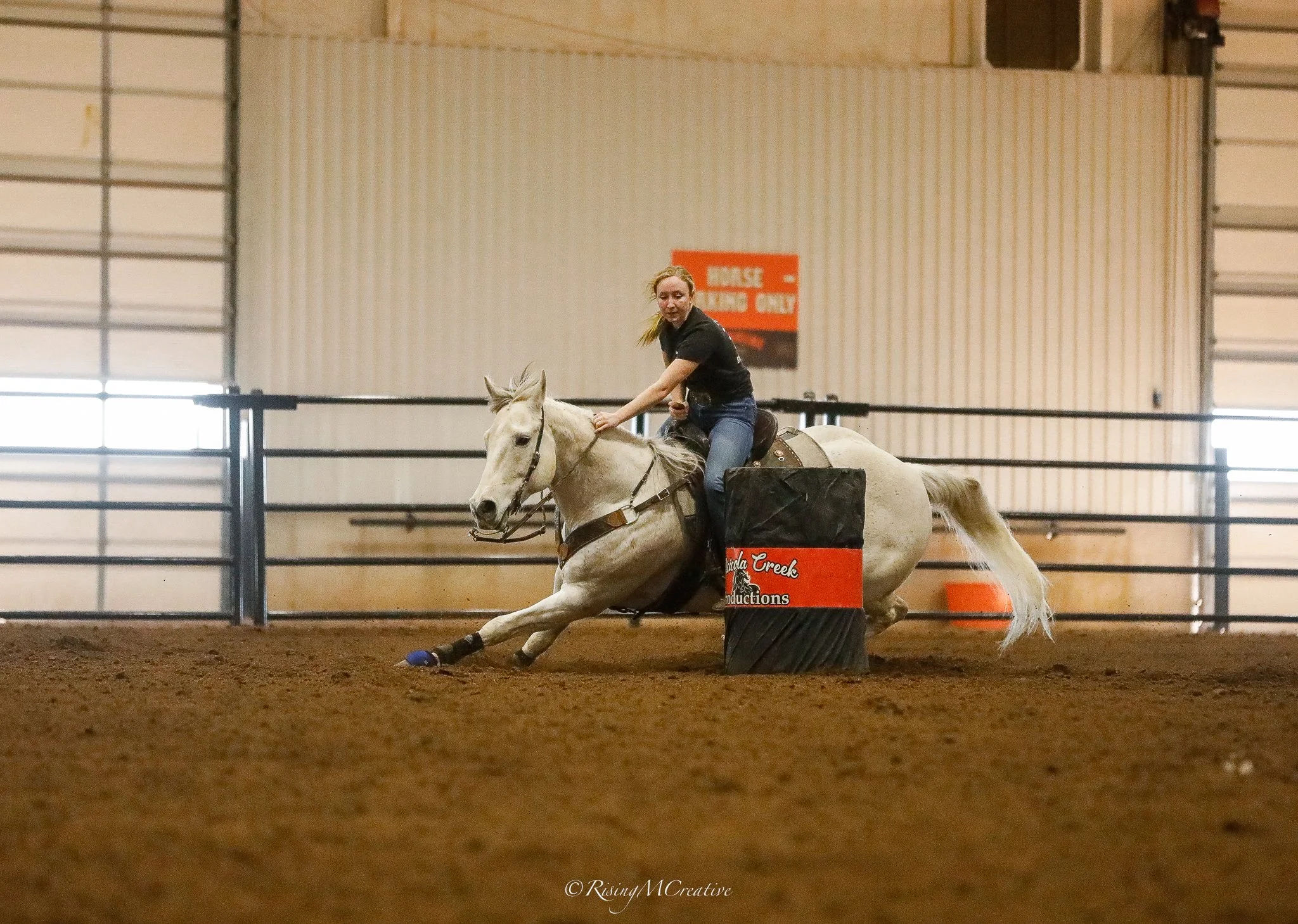 A woman riding a white horse in an indoor arena during a barrel racing event, with orange barrels and a sign in the background.