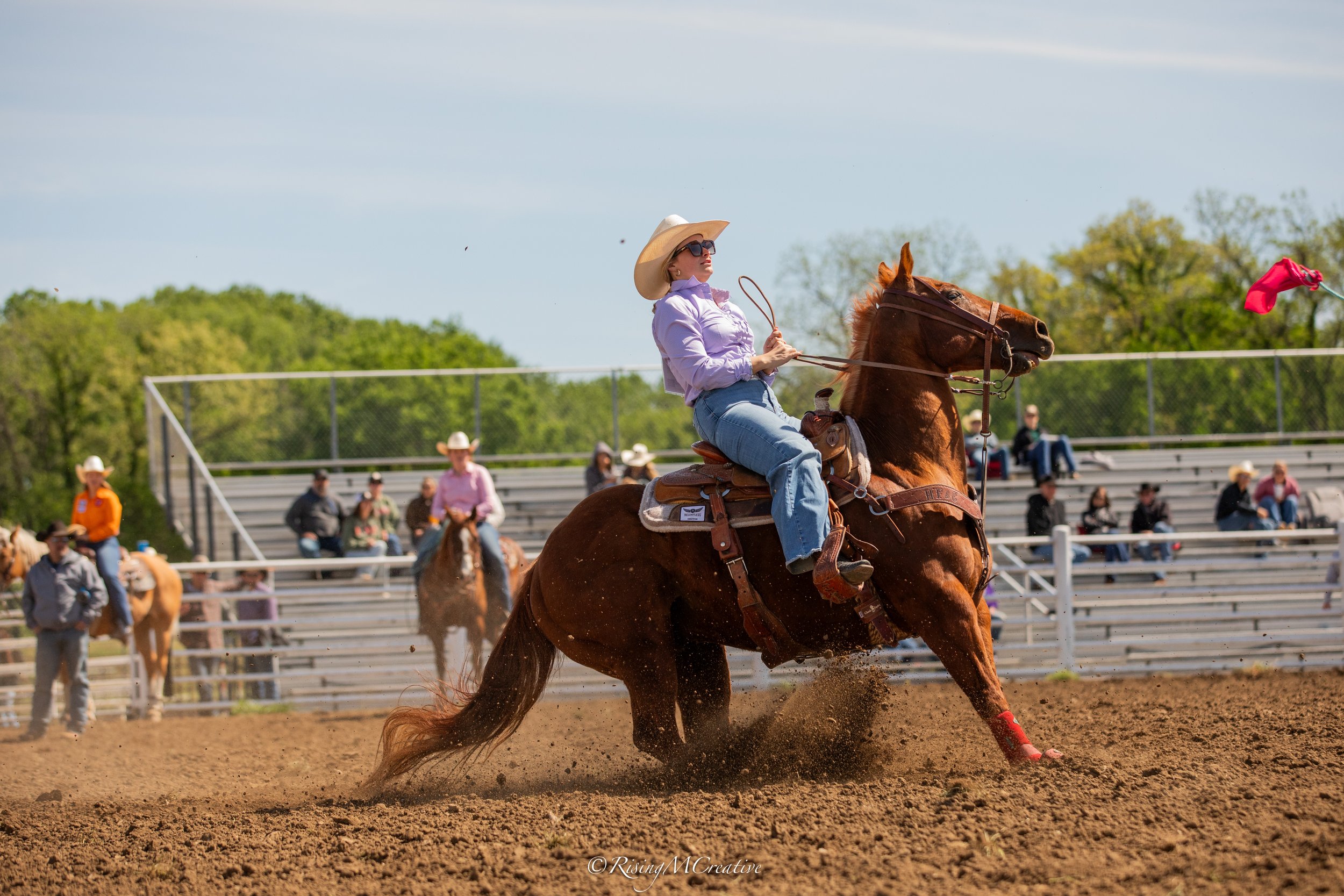 A woman riding a galloping brown horse on a dirt rodeo arena, wearing a cowboy hat, sunglasses, and a purple shirt. Spectators are seated on bleachers in the background, with some wearing cowboy hats.