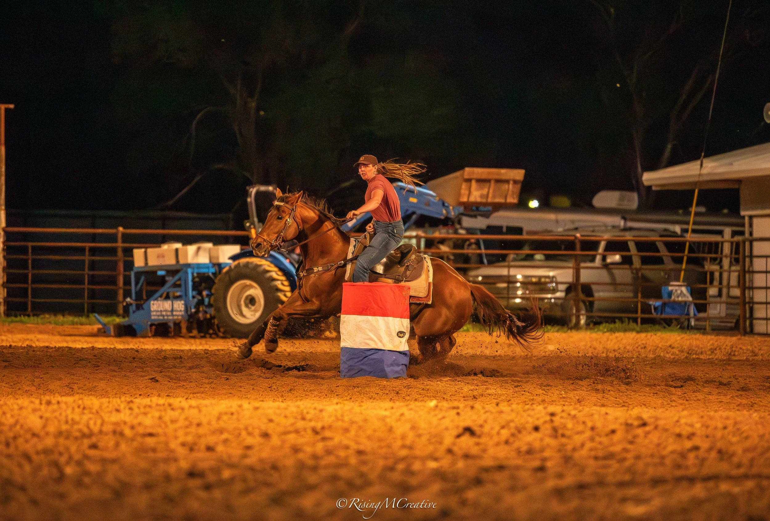 A woman riding a horse during a nighttime rodeo event. She is wearing a baseball cap, a red shirt, and jeans, and is in the middle of a riding maneuver on the horse, which is kicking up dirt. The scene is illuminated by artificial lights, with equipm