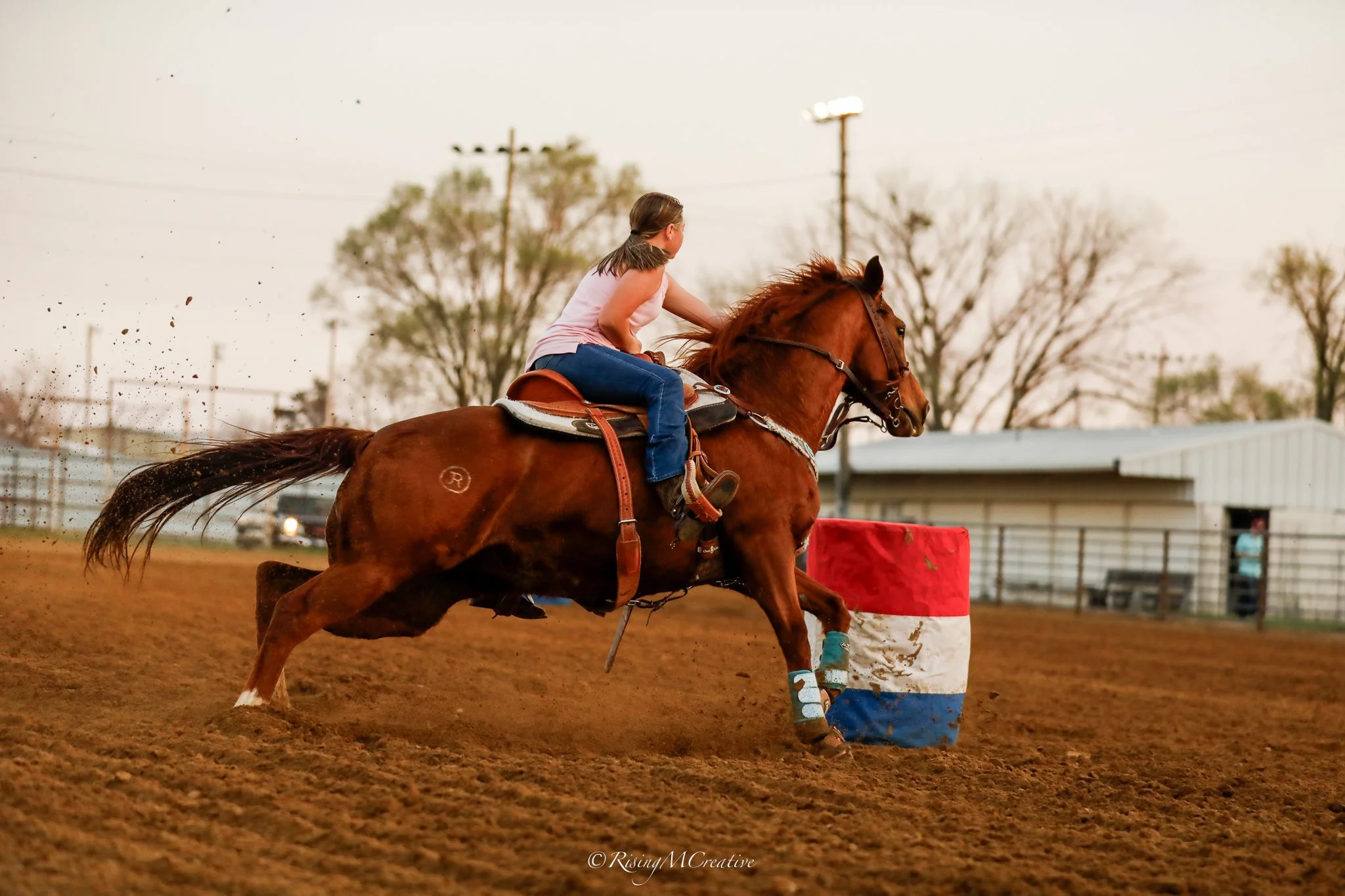 A girl riding a horse around a dirt race track during a rodeo event, with barriers and a barn in the background.