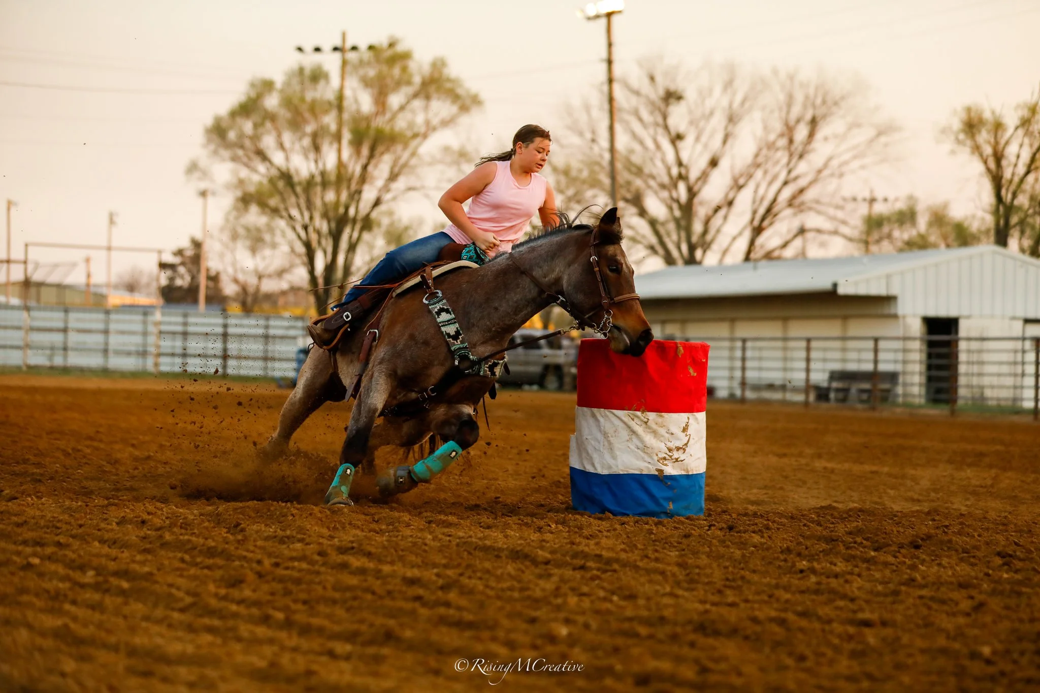 A young woman riding a horse during a barrel racing event on a dirt track as she navigates around a colored barrel.