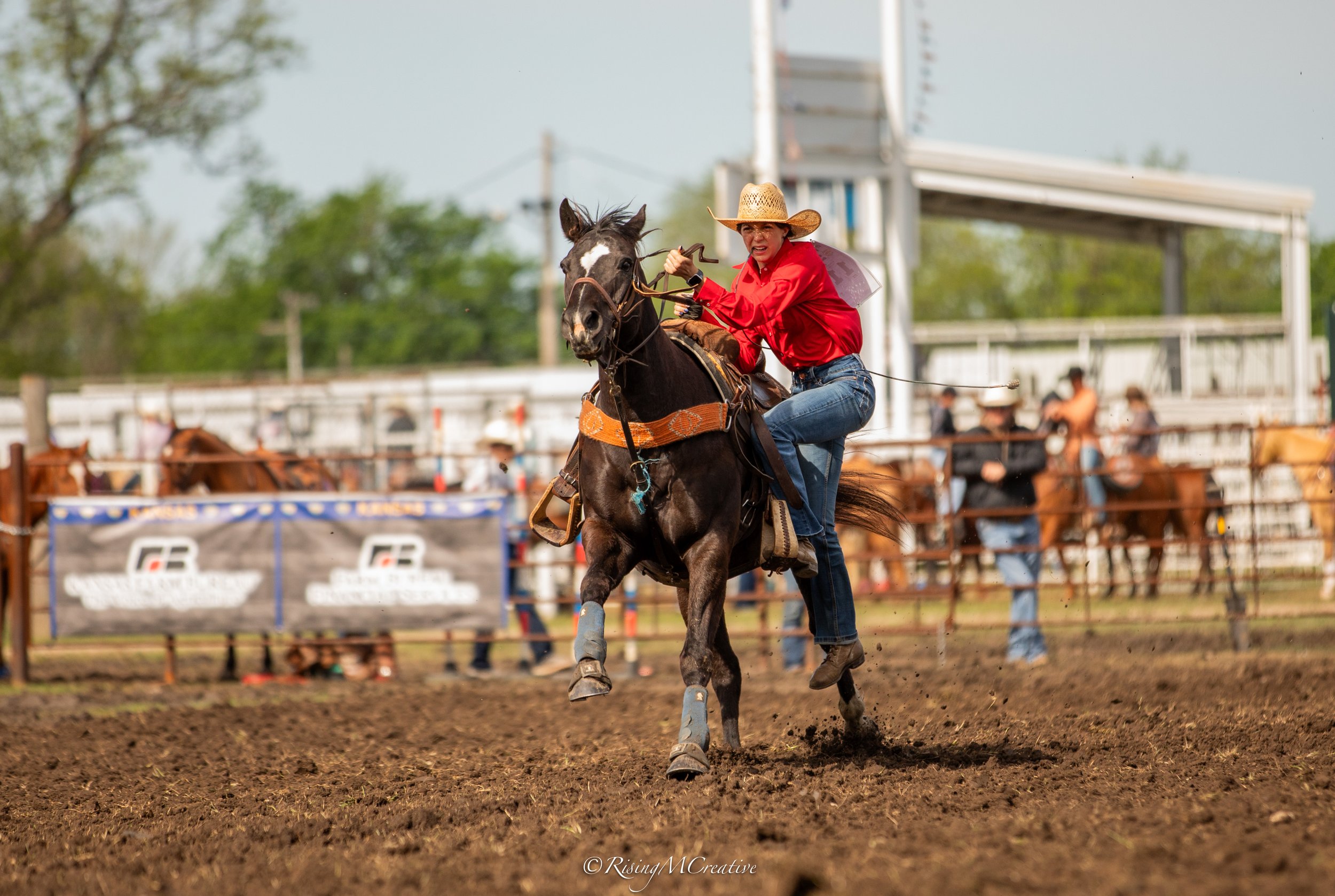 A female rider wearing a straw hat, red shirt, and jeans riding a galloping horse in a rodeo arena with people and banners in the background.