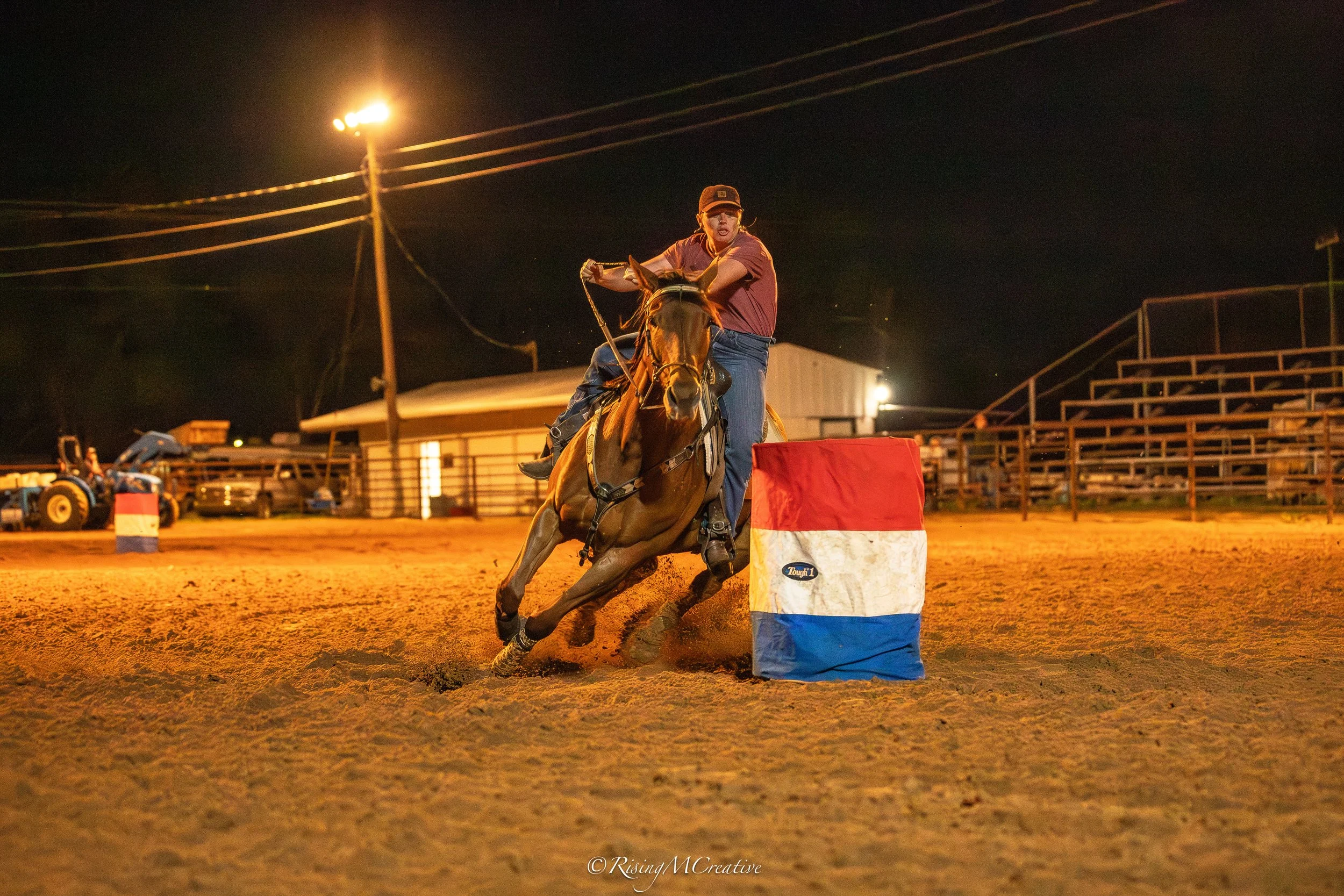 A person riding a horse in a rodeo arena at night, navigating a barrel while the crowd watches. The rider is wearing a brown cap and maroon shirt, and the horse is in motion on a dirt track. Bright lights illuminate the scene.
