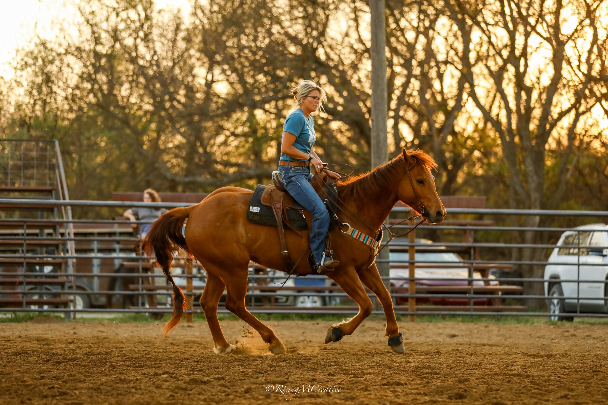 A woman riding a brown horse in an outdoor arena during sunset, with trees and fencing in the background.