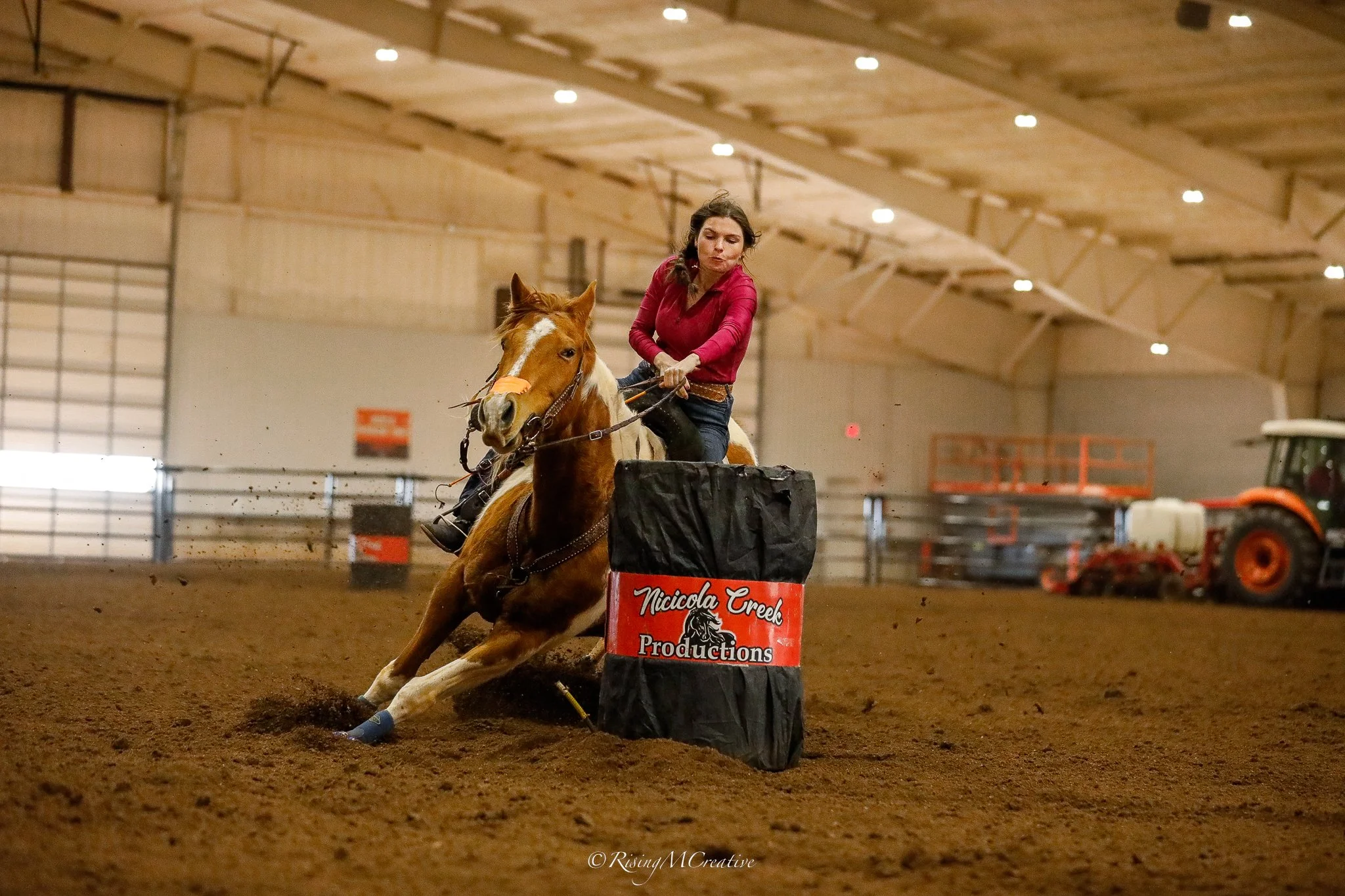 A woman riding a horse in an indoor arena, navigating between obstacles during a barrel racing event. The arena has a dirt floor and is well-lit with wooden and metal structures.