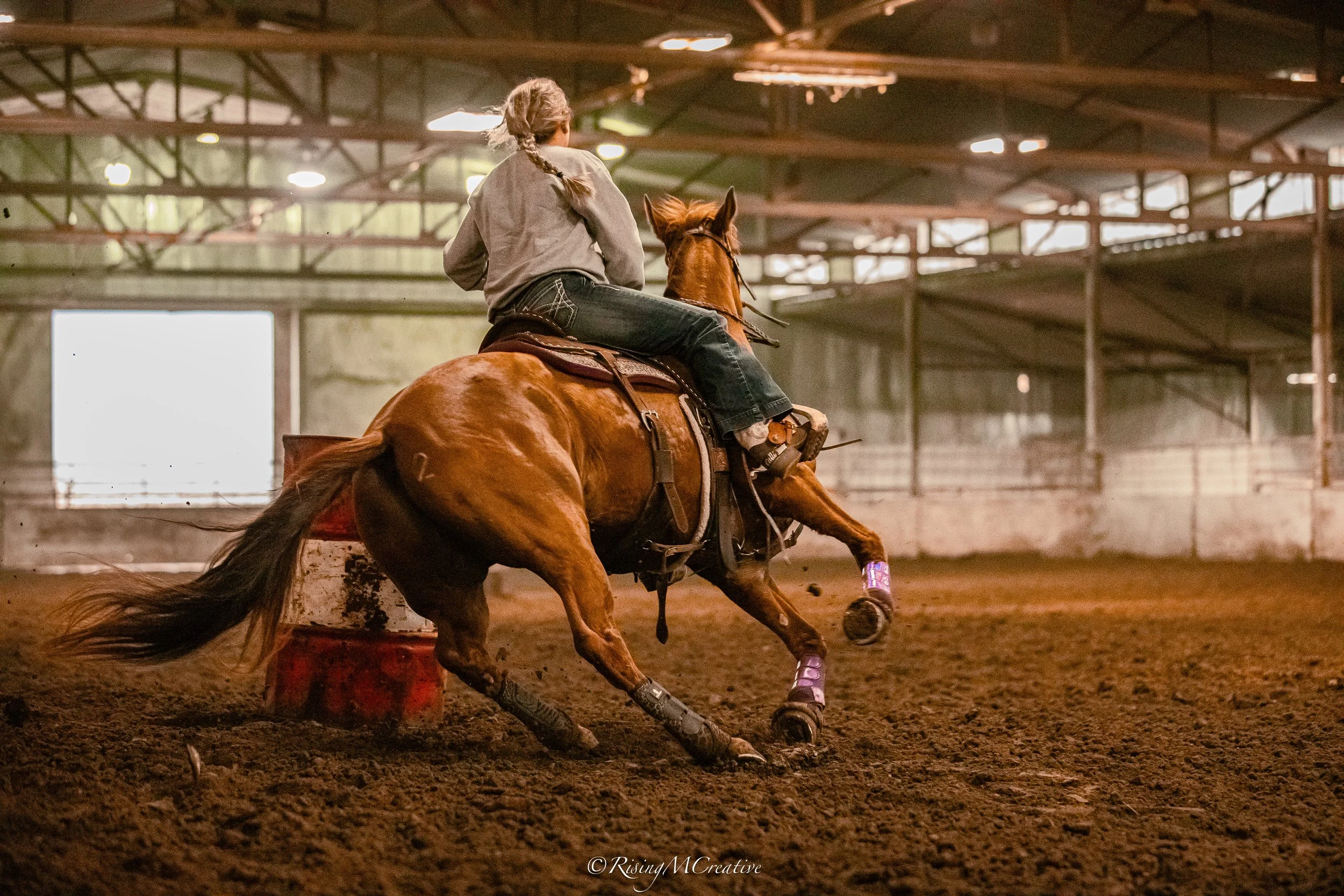 A rider on a galloping horse inside an indoor arena, with a dirt floor and large windows letting in natural light.