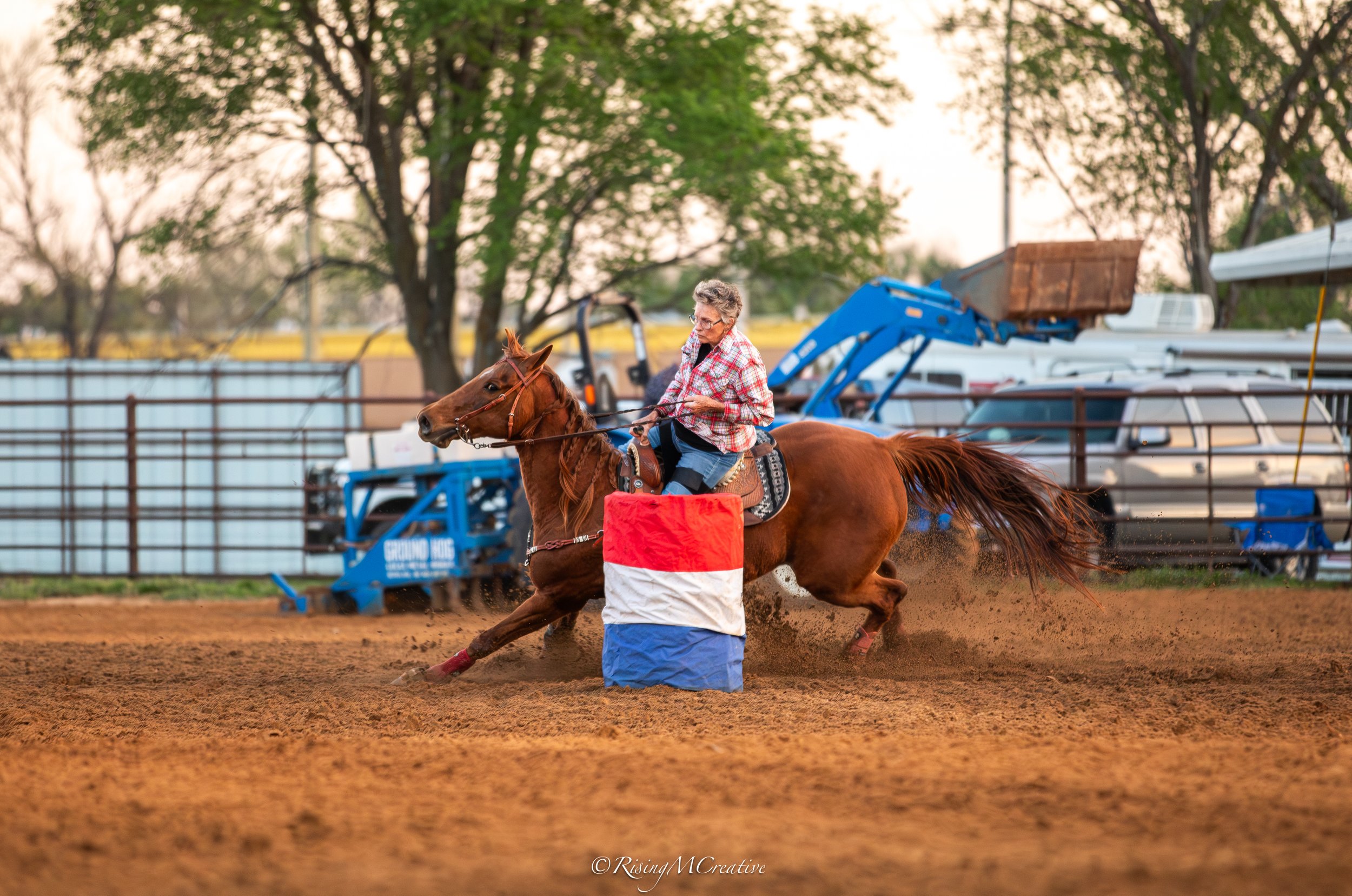 A woman riding a galloping horse in an outdoor arena surrounded by trees, with some equipment and vehicles in the background during sunset.