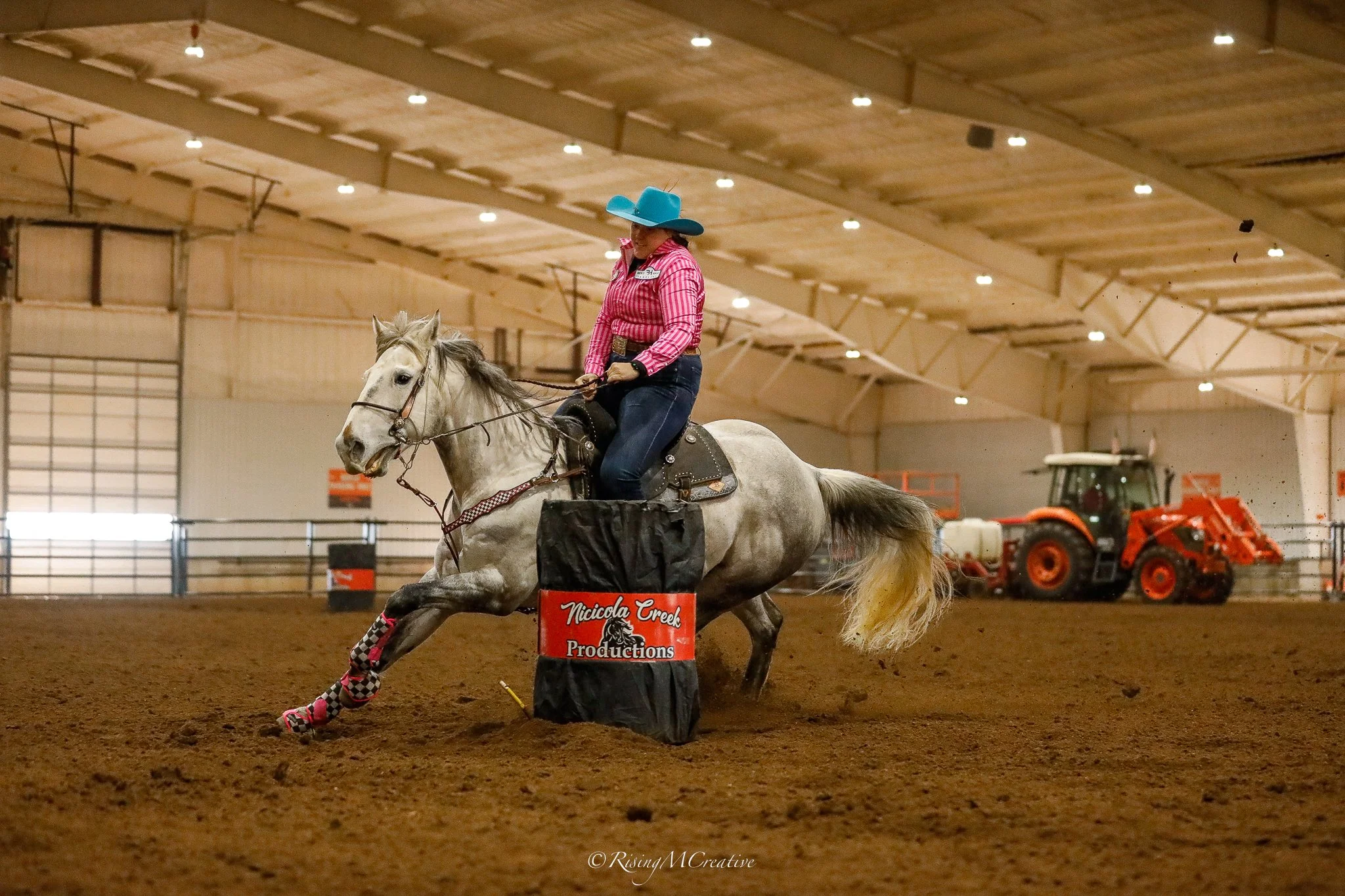 A woman riding a white horse inside an indoor equestrian arena, wearing a cowboy hat, pink striped shirt, and checkered leg wraps, navigating around an obstacle labeled 'Niccola Creek Productions'.